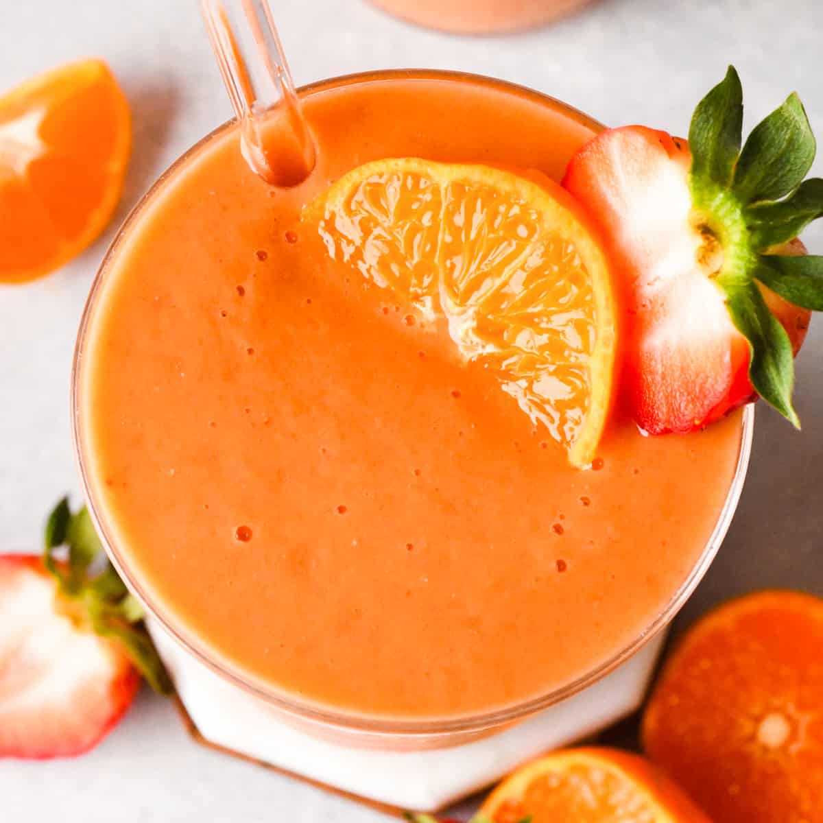Tall glass of orange-colored carrot fruit smoothie garnished with a strawberry and an orange slice, with a straw in the glass and fresh fruit in the background.
