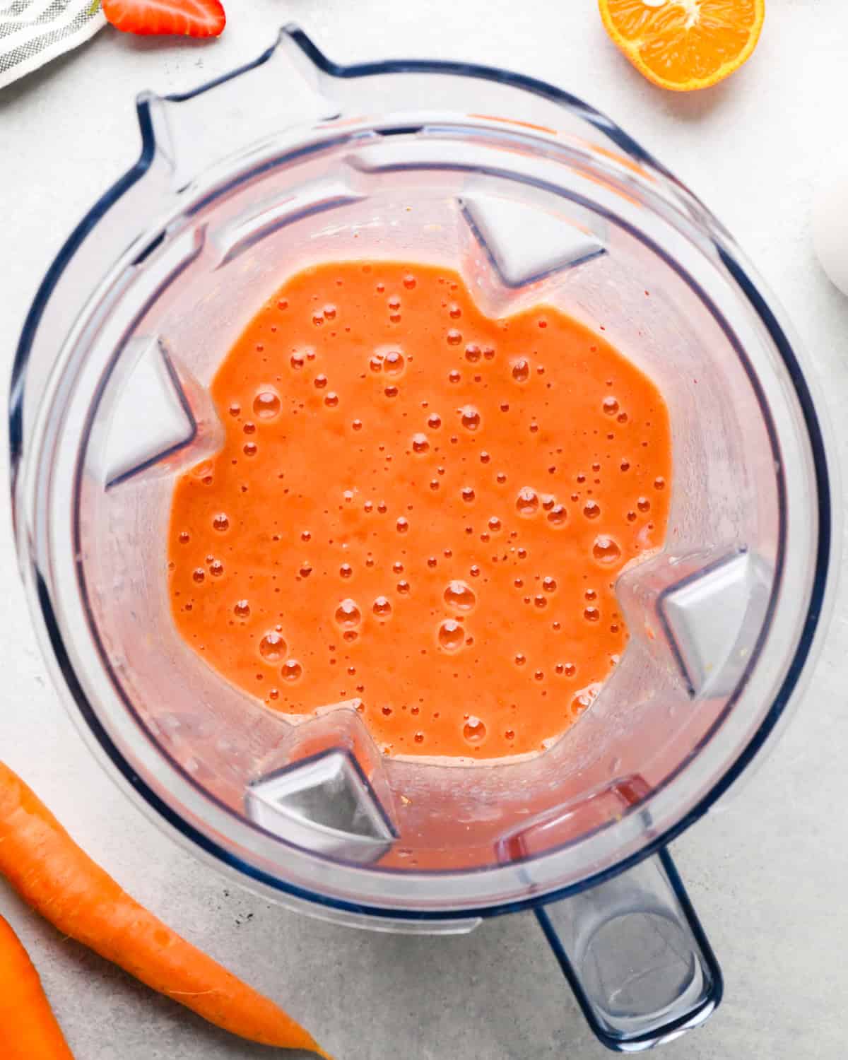 Making carrot fruit smoothie: overhead view of milk, oranges, strawberries, bananas, honey, and carrots sitting in a blender and blended together. Carrots and fruit slices are sitting near the blender on a light surface.