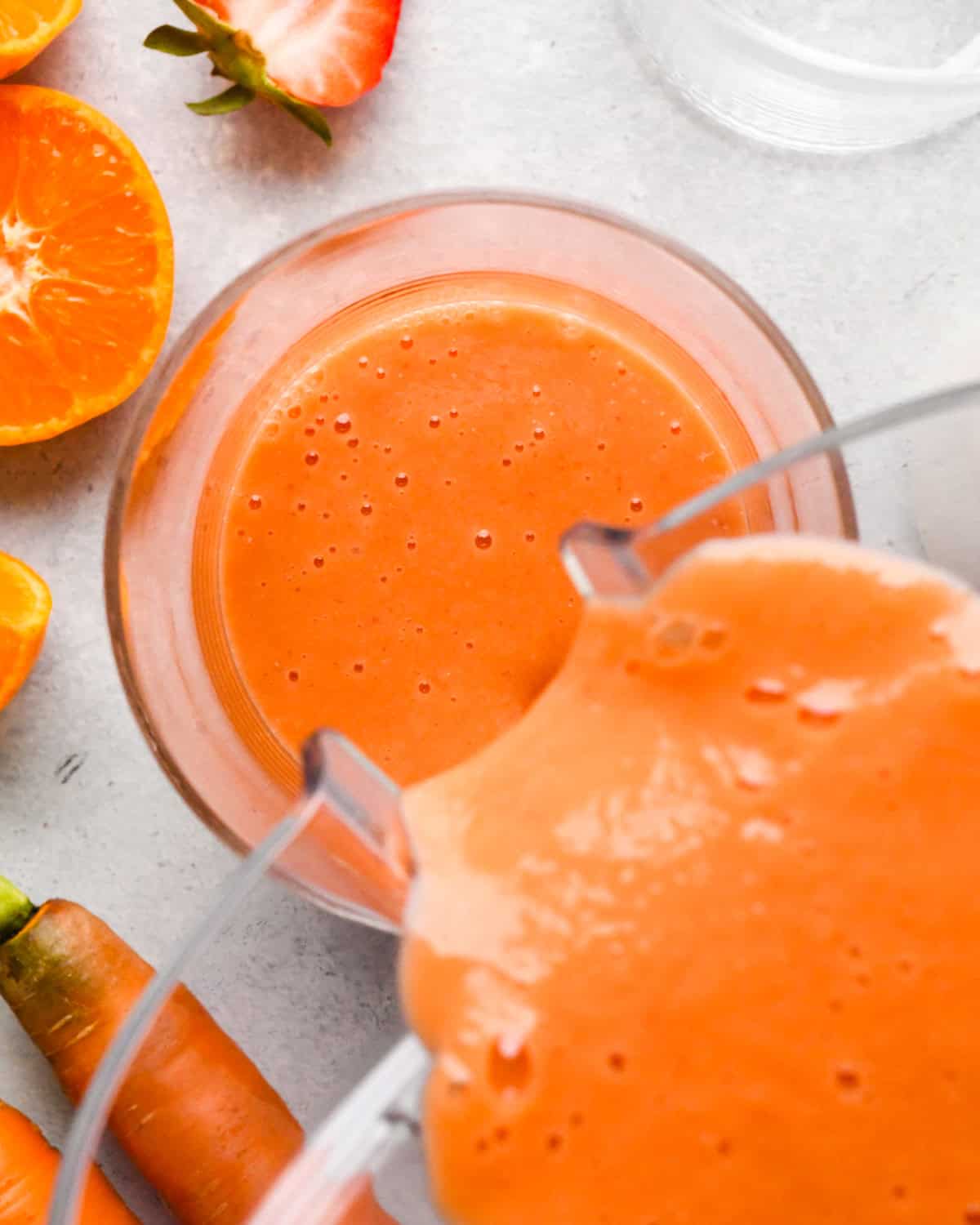 Making carrot fruit smoothie: overhead view of completed smoothie being poured into a tall clear glass on a light surface.