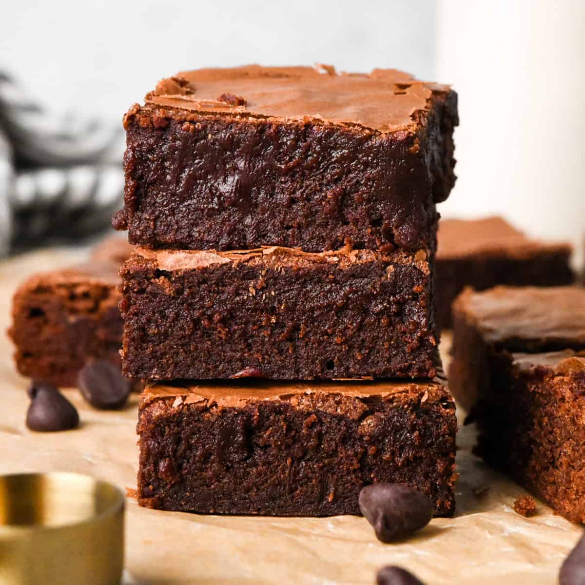 Stack of rich, fudgy gluten-free brownies with a crackly top, shown close-up on parchment paper with chocolate chips nearby.