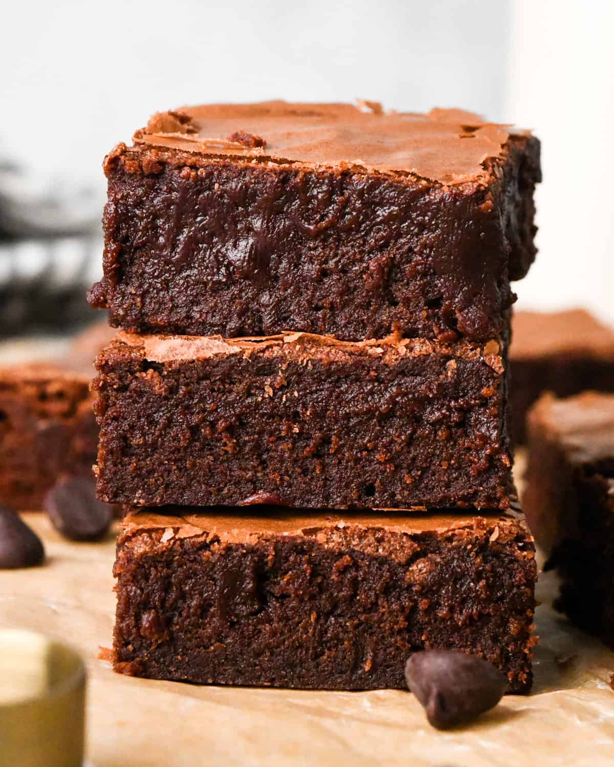 Stack of rich, fudgy gluten-free brownies with a crackly top, shown close-up on parchment paper with chocolate chips nearby.