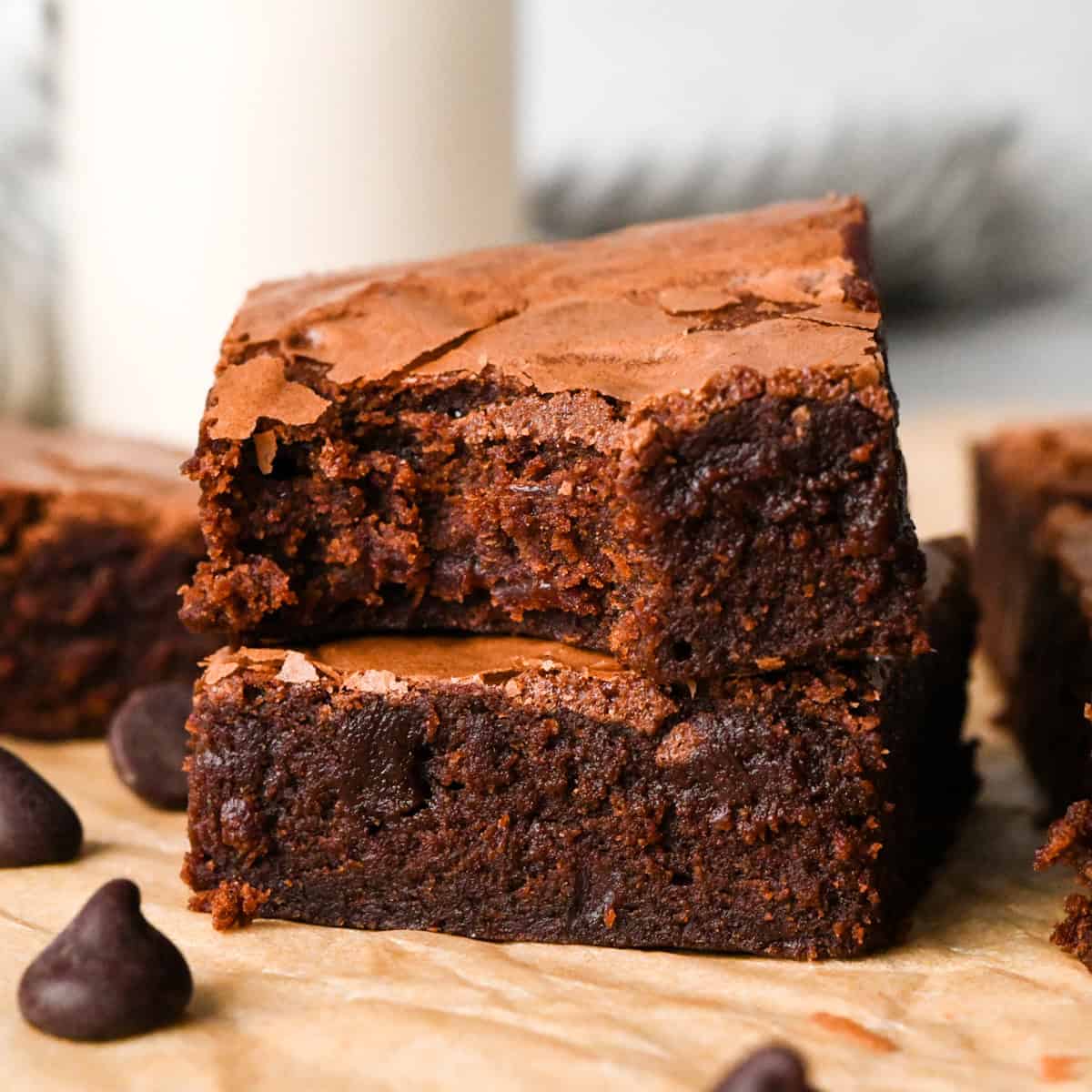 Stack of rich, fudgy gluten-free brownies with a crackly top, shown close-up on parchment paper with chocolate chips nearby and a bite taken out of the top brownie.