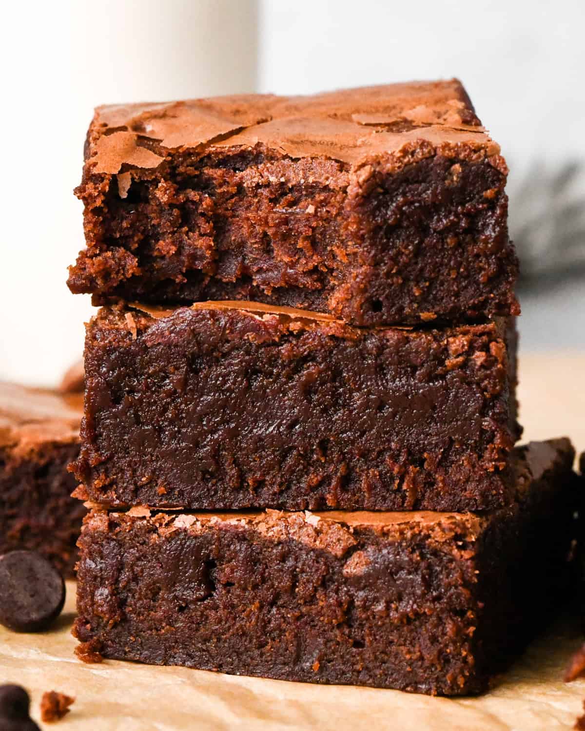 Stack of rich, fudgy gluten-free brownies with a crackly top, shown close-up on parchment paper with chocolate chips nearby and a bite taken out of the top brownie.