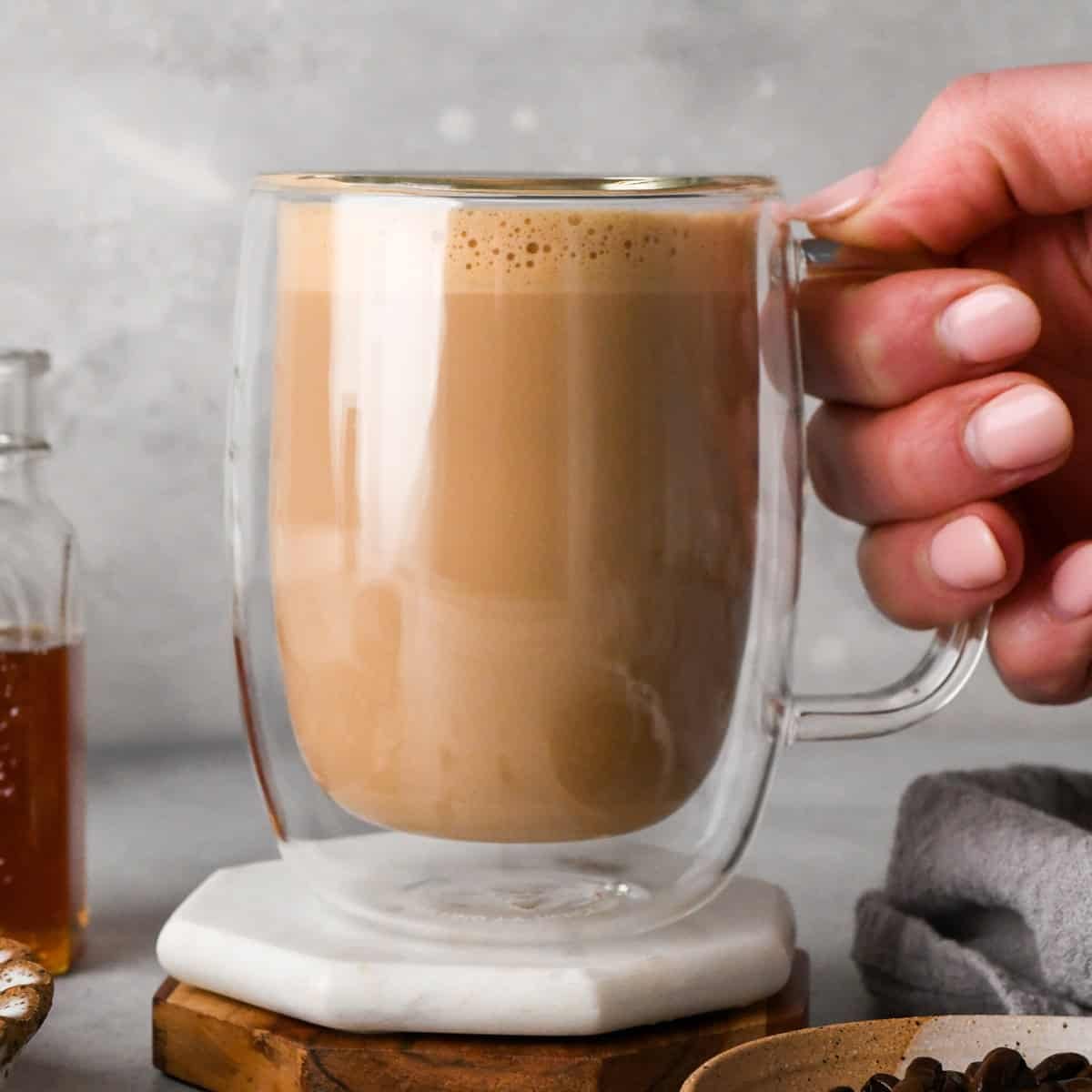 a hand holding a clear mug with a homemade latte inside, there is a glass jar of honey in the background and a bowl of coffee beans in the foreground