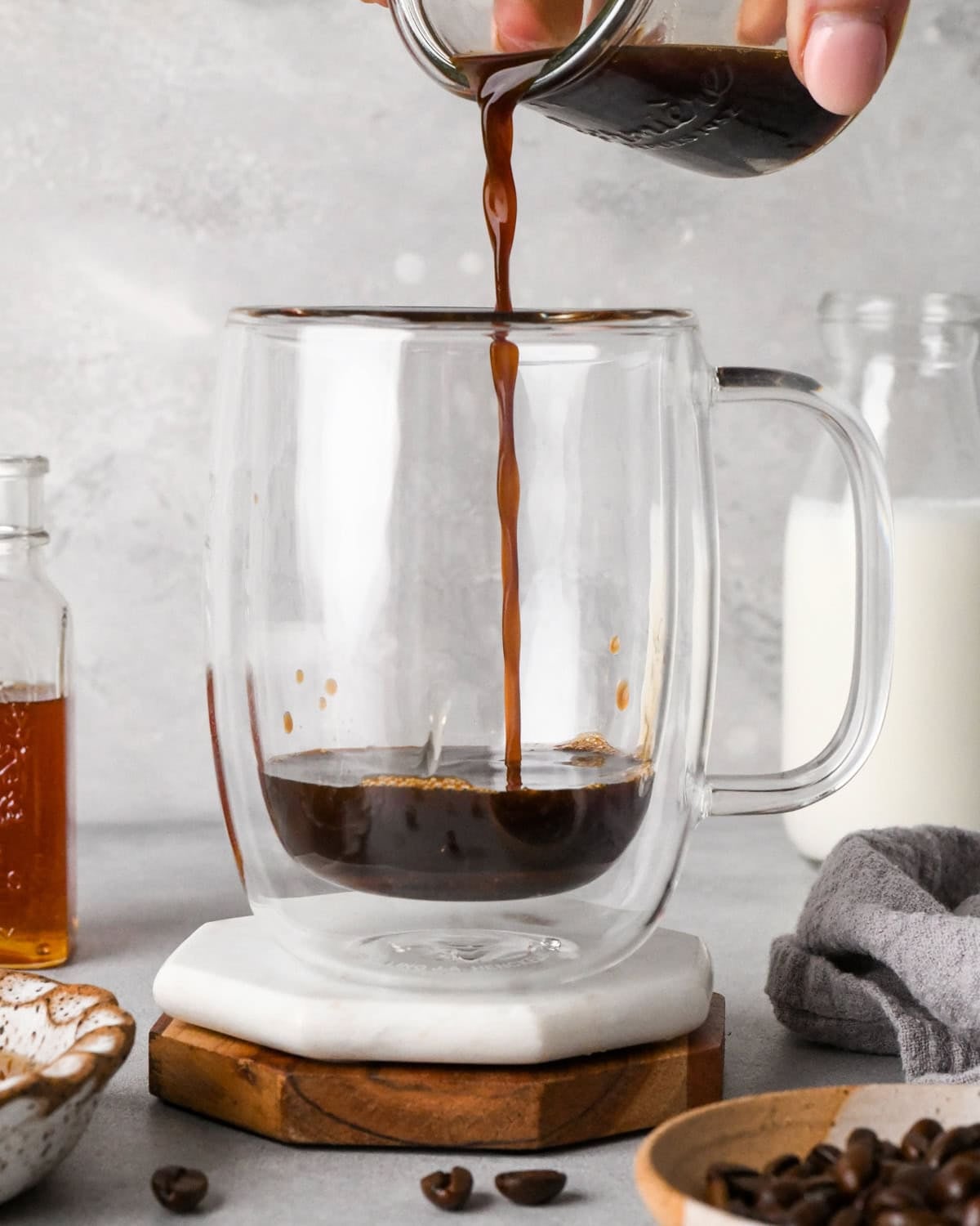 How to make a latte: front-view photo of espresso being added to the honey in a clear coffee mug. The mug is sitting on a white coaster and surrounded by additional ingredients like coffee beans, honey, and milk.