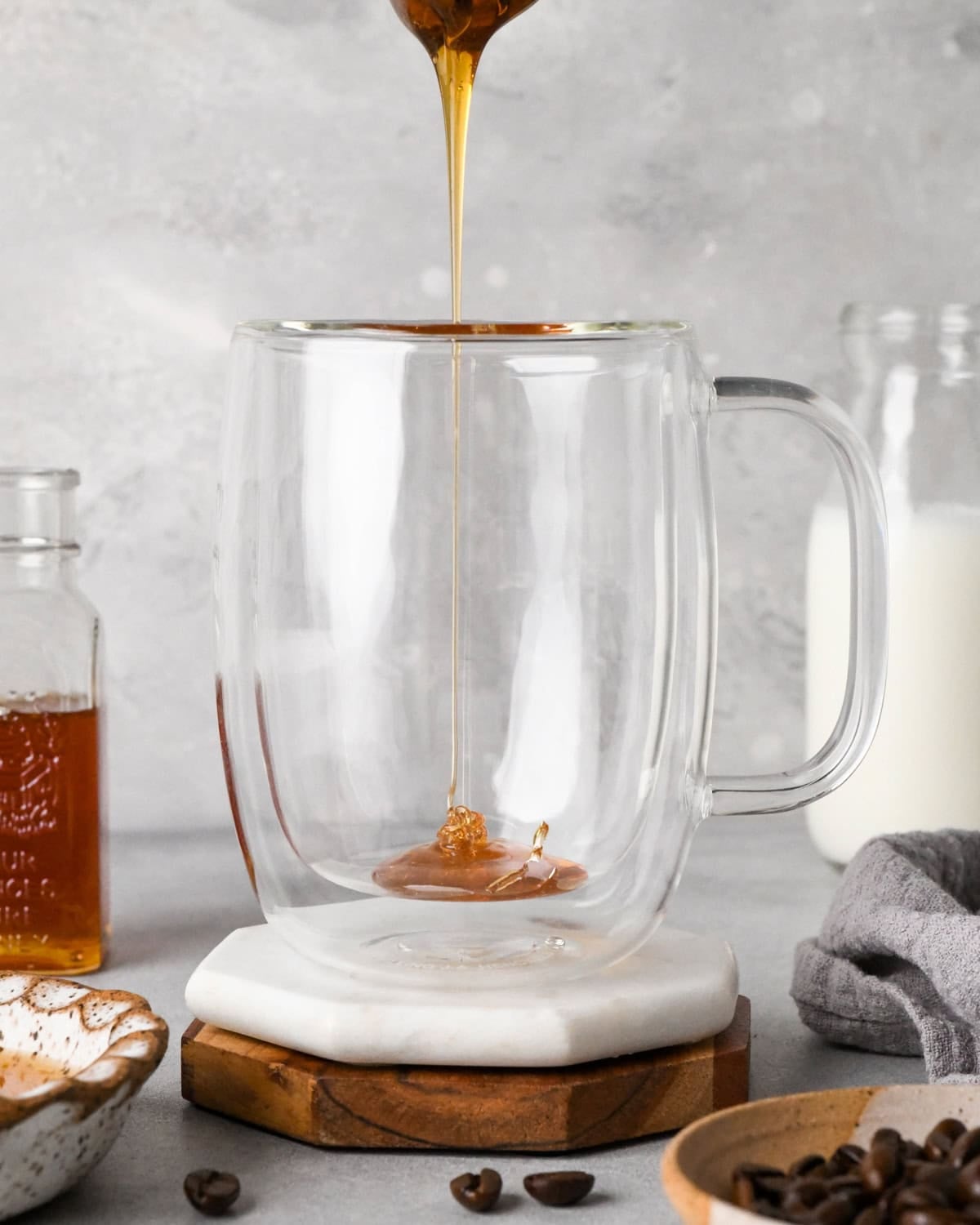 How to make a latte: front-view photo of honey being poured into a clear coffee mug on a white coaster.