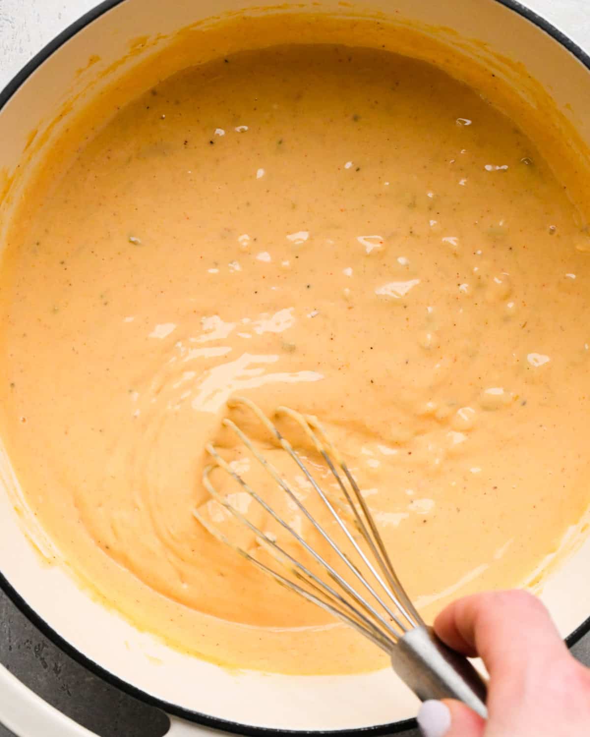 Making homemade queso: overhead photo of green chiles and spices mixed into the simmering sauce in a large white pot and whisked together. We see that the sauce has started to thicken.