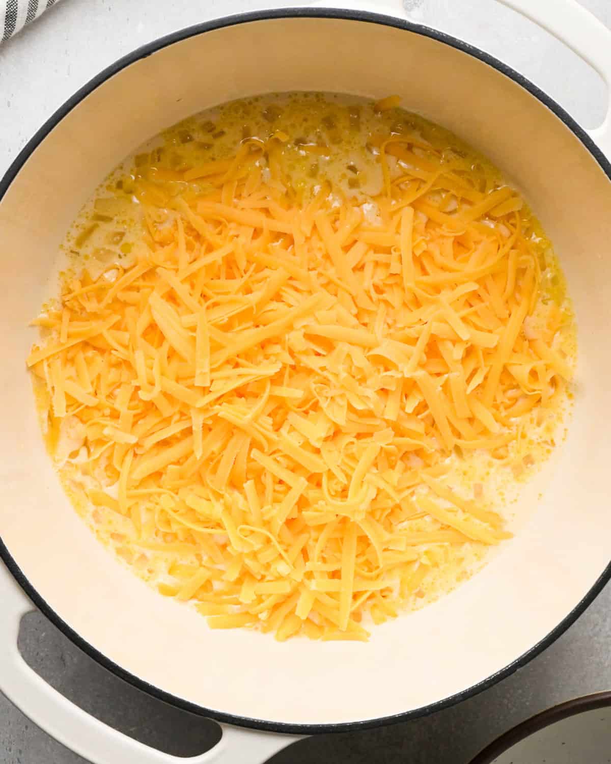 Making homemade queso: overhead photo of shredded cheese being added to large white pot with the simmering sauce.