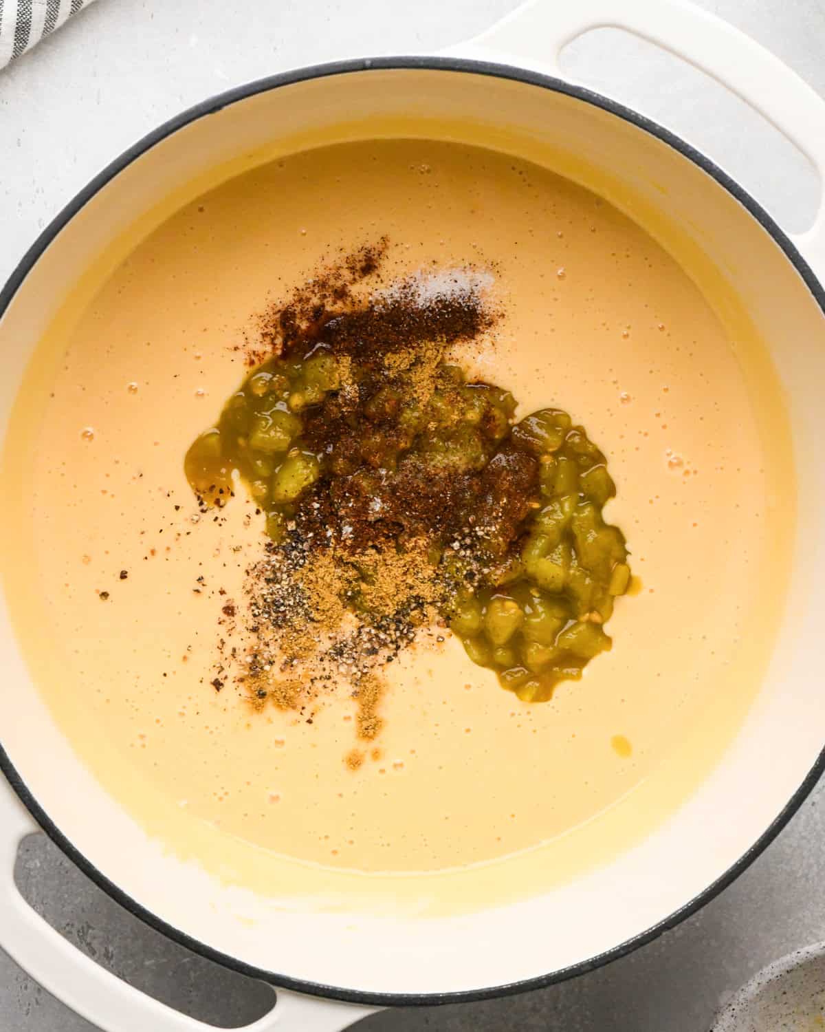 Making homemade queso: overhead photo of green chiles and spices being added to the simmering sauce in a large white pot.