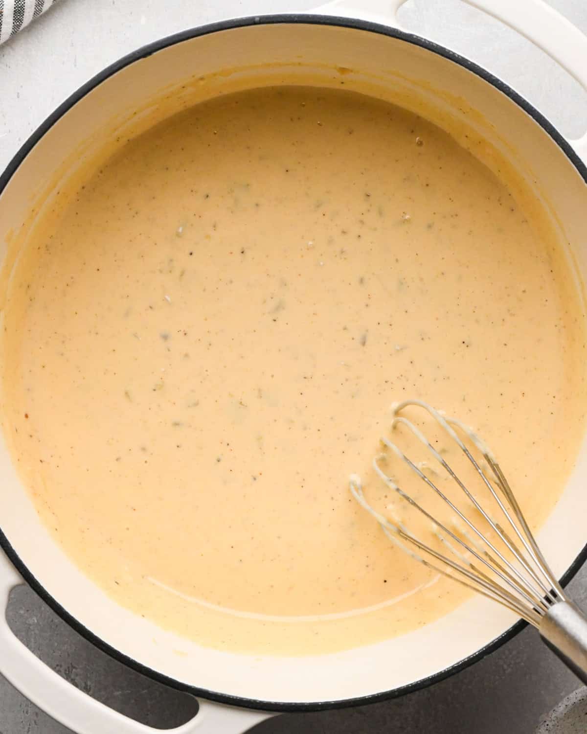 Making homemade queso: overhead photo of green chiles and spices mixed into the simmering sauce in a large white pot and whisked together.