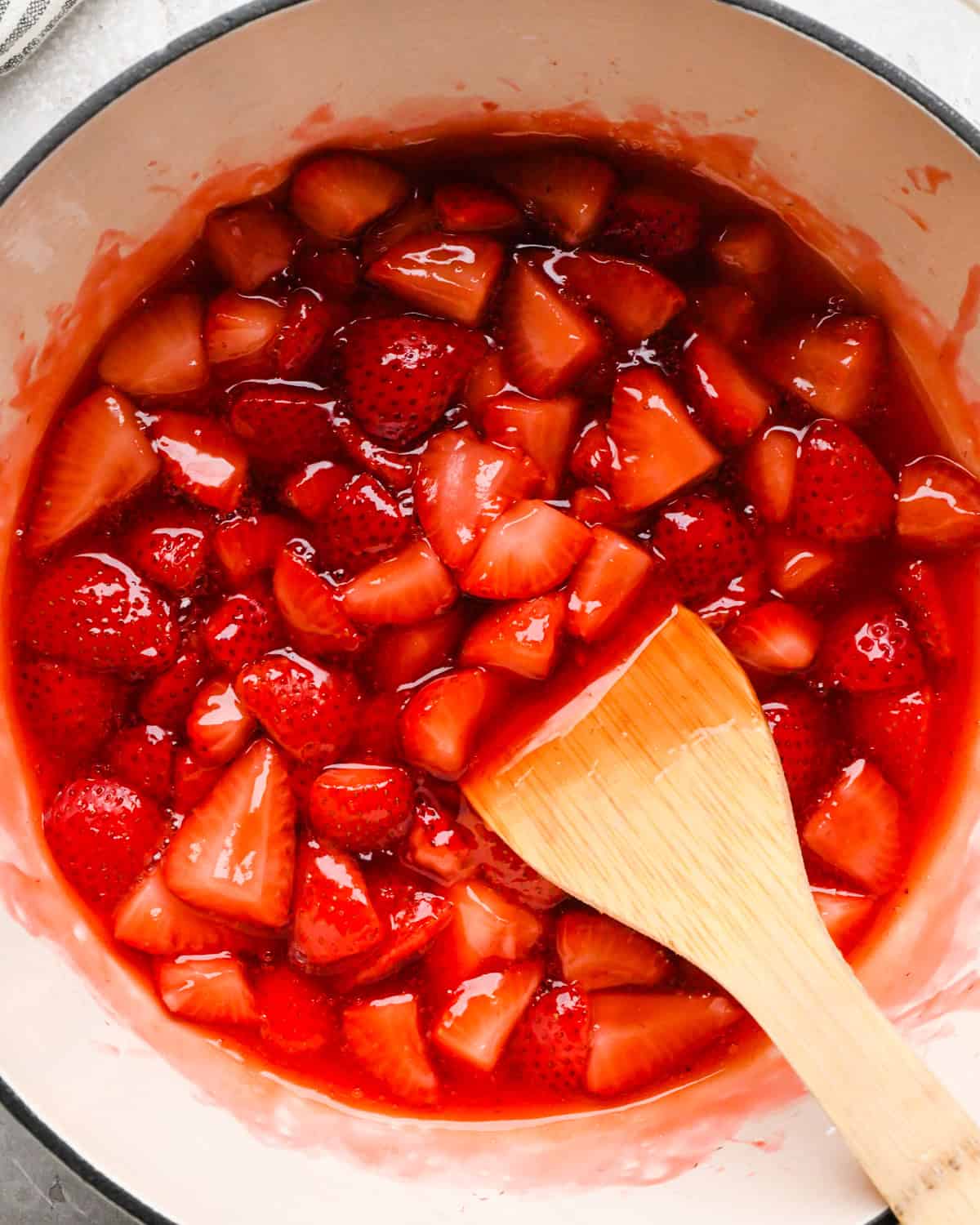 Making homemade strawberry sauce: overhead photo of strawberries, sugar, lemon juice, and cornstarch/water mixture simmering together in a large white pot. A wooden spoon is stirring the sauce.