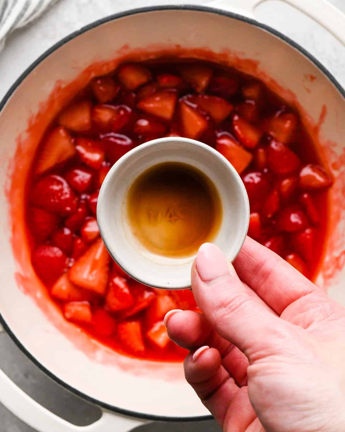 Making homemade strawberry sauce: overhead photo a hand holding a small bowl of vanilla ready to pour it into the strawberry sauce.
