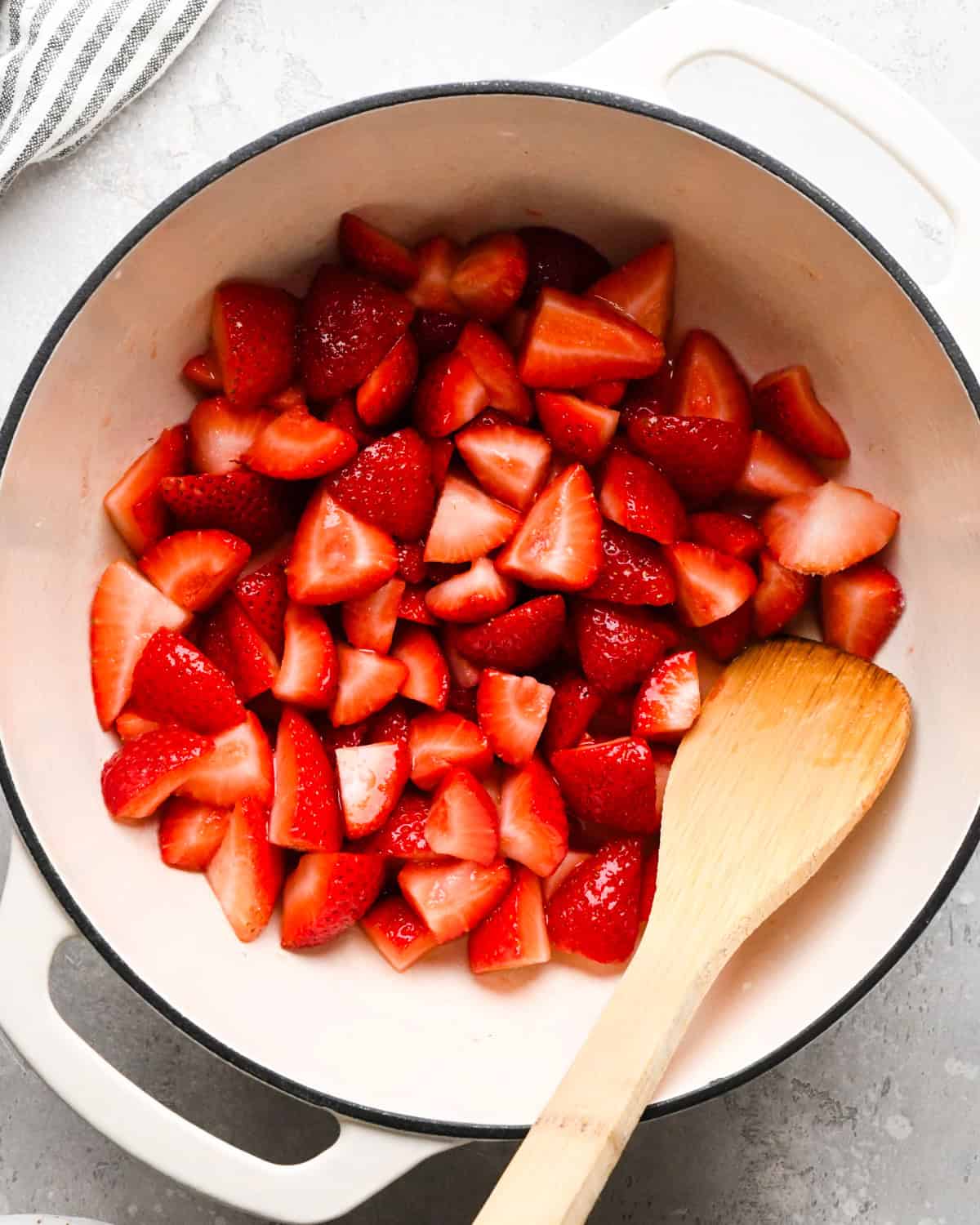 Making homemade strawberry sauce: overhead photo of strawberries, sugar, lemon juice, and cornstarch/water mixture in a large white pot being mixed together with a wooden spoon.