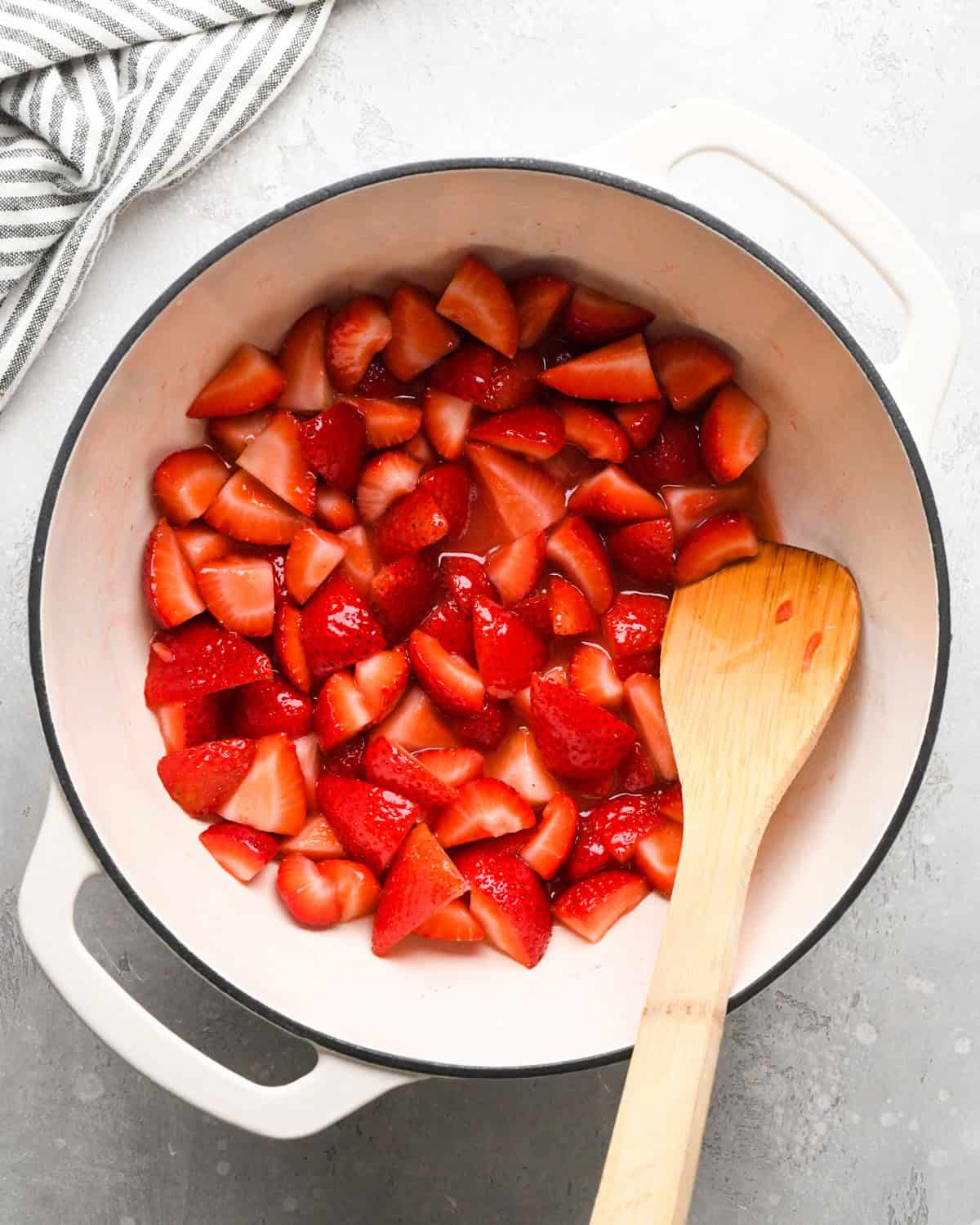 Making homemade strawberry sauce: overhead photo of strawberries, sugar, lemon juice, and cornstarch/water mixture simmering together in a large white pot. A wooden spoon is sitting in the pot.