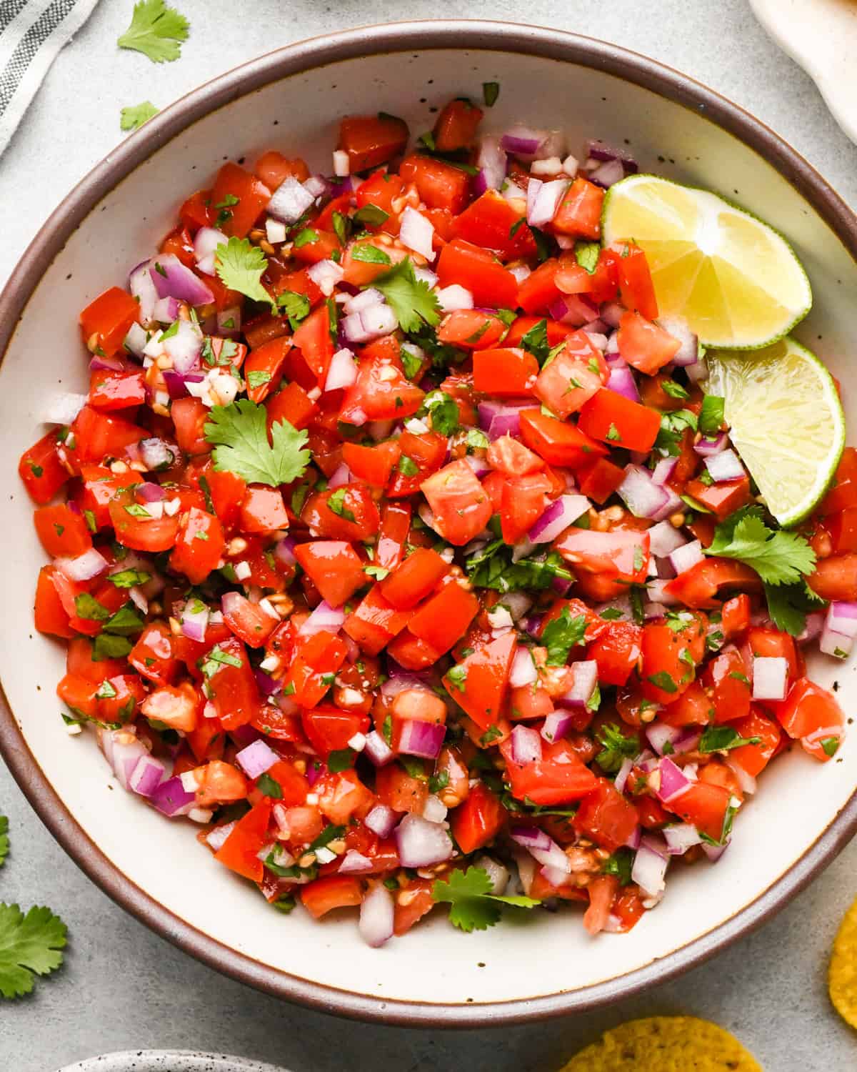 Making pico de Gallo: overhead photo of completed pico de Gallo in a large white bowl with lime wedges in the bowl and tortilla chips nearby on a light surface.