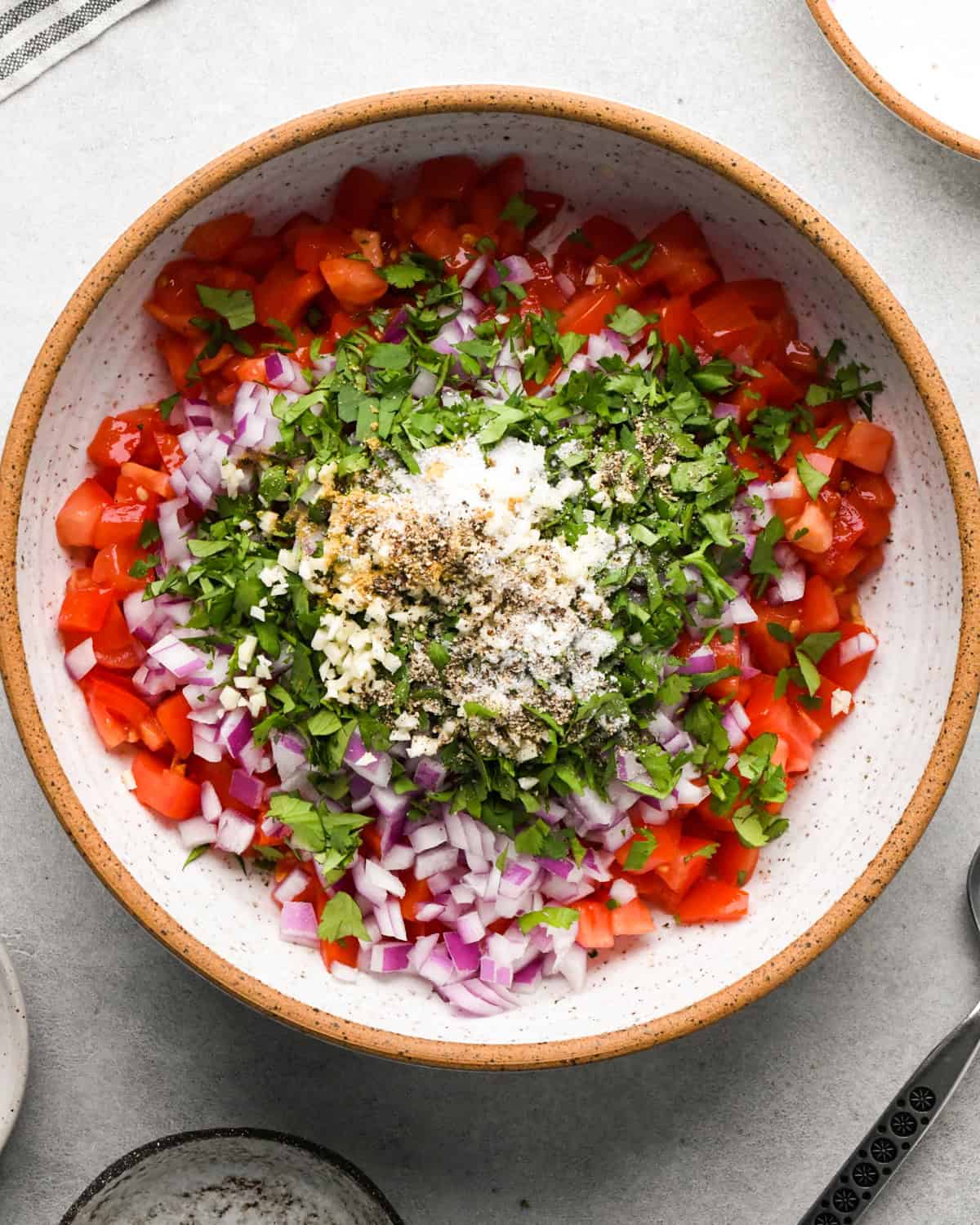 Making pico de Gallo: overhead photo of a white speckled bowl filled with diced tomatoes, cilantro, minced garlic, and red onion. Spices including salt, pepper, and cumin are added on top.