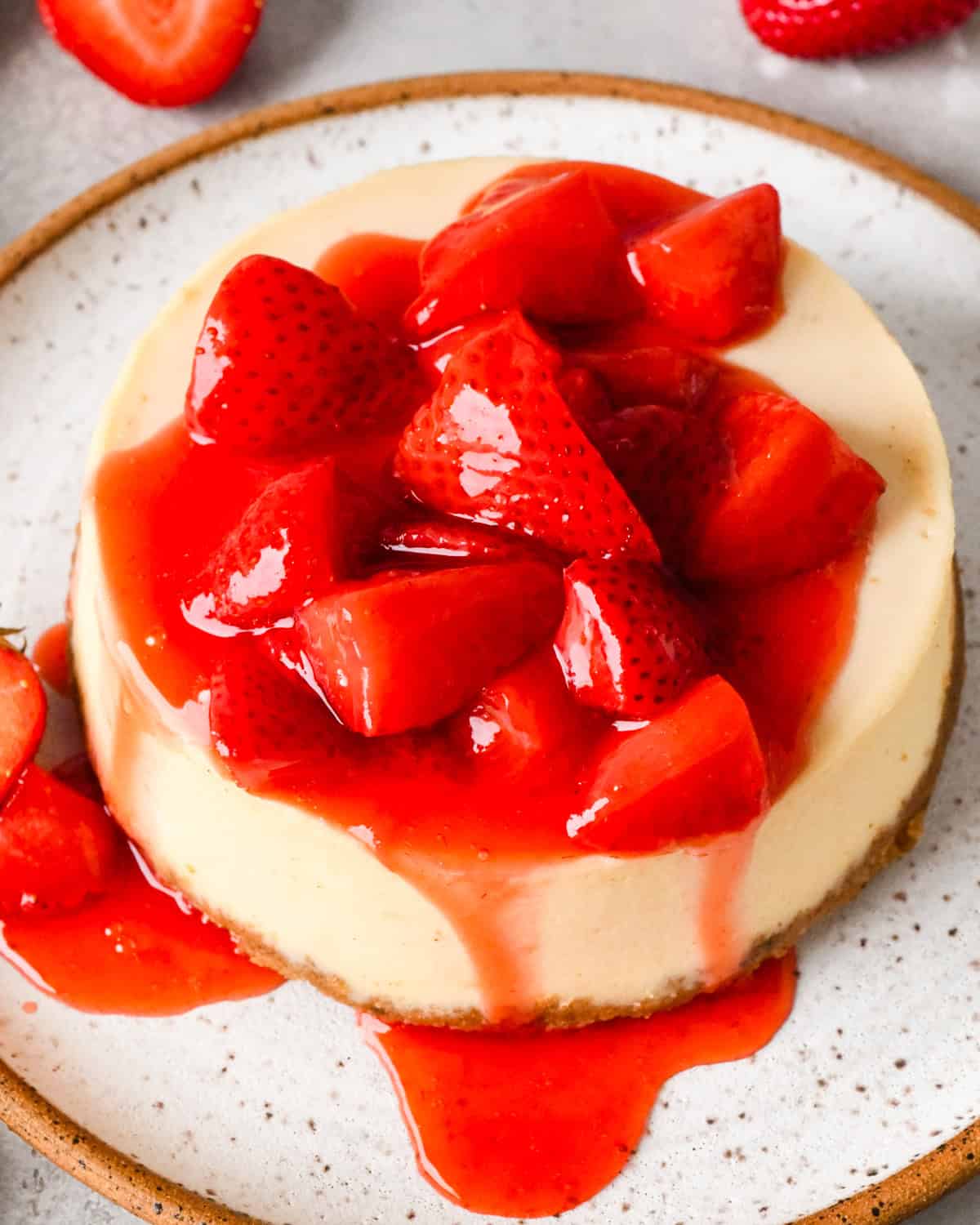 Close-up of a cheesecake topped with homemade strawberry sauce and fresh strawberry pieces, with red sauce dripping over the sides.