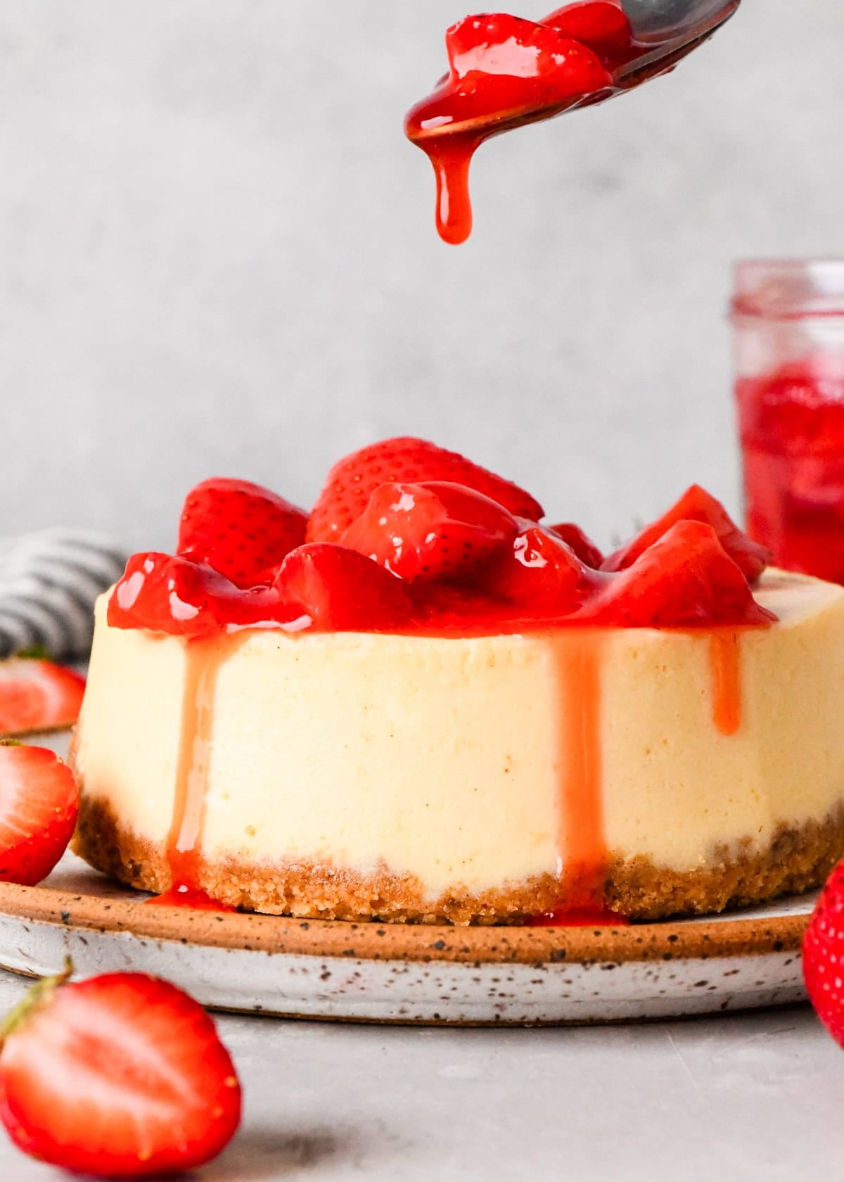 Close-up of a cheesecake topped with homemade strawberry sauce and fresh strawberry pieces, with red sauce dripping over the sides. A spoon is pouring additional sauce onto the cheesecake.