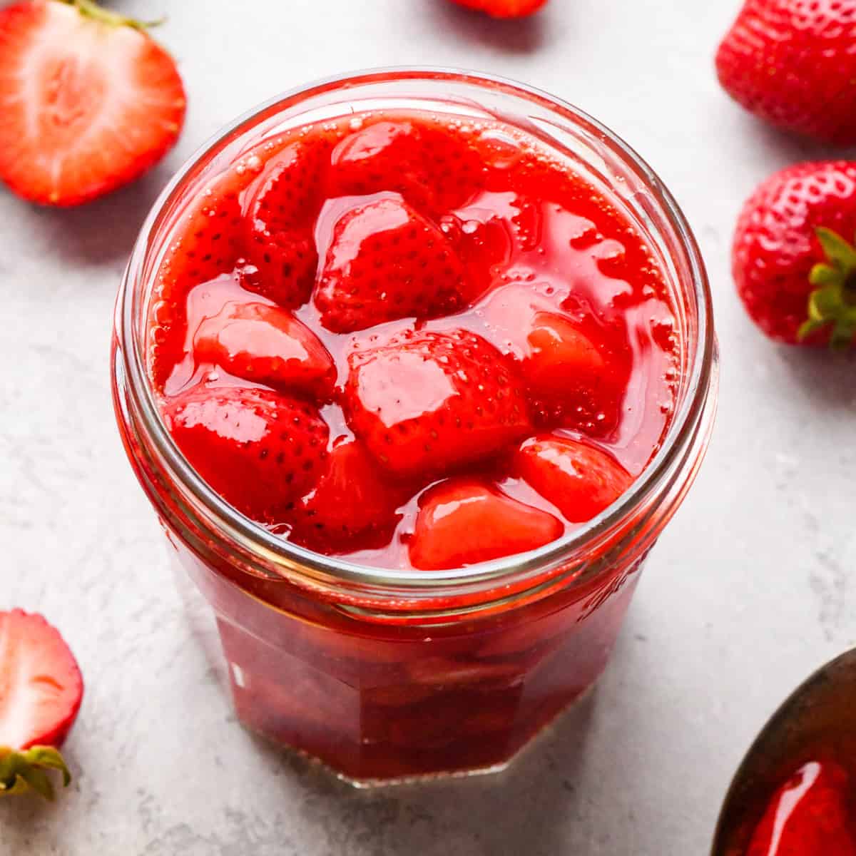 Overhead photo of a clear jar filled with bright red homemade strawberry sauce, with extra strawberries surrounding the jar on a light surface.