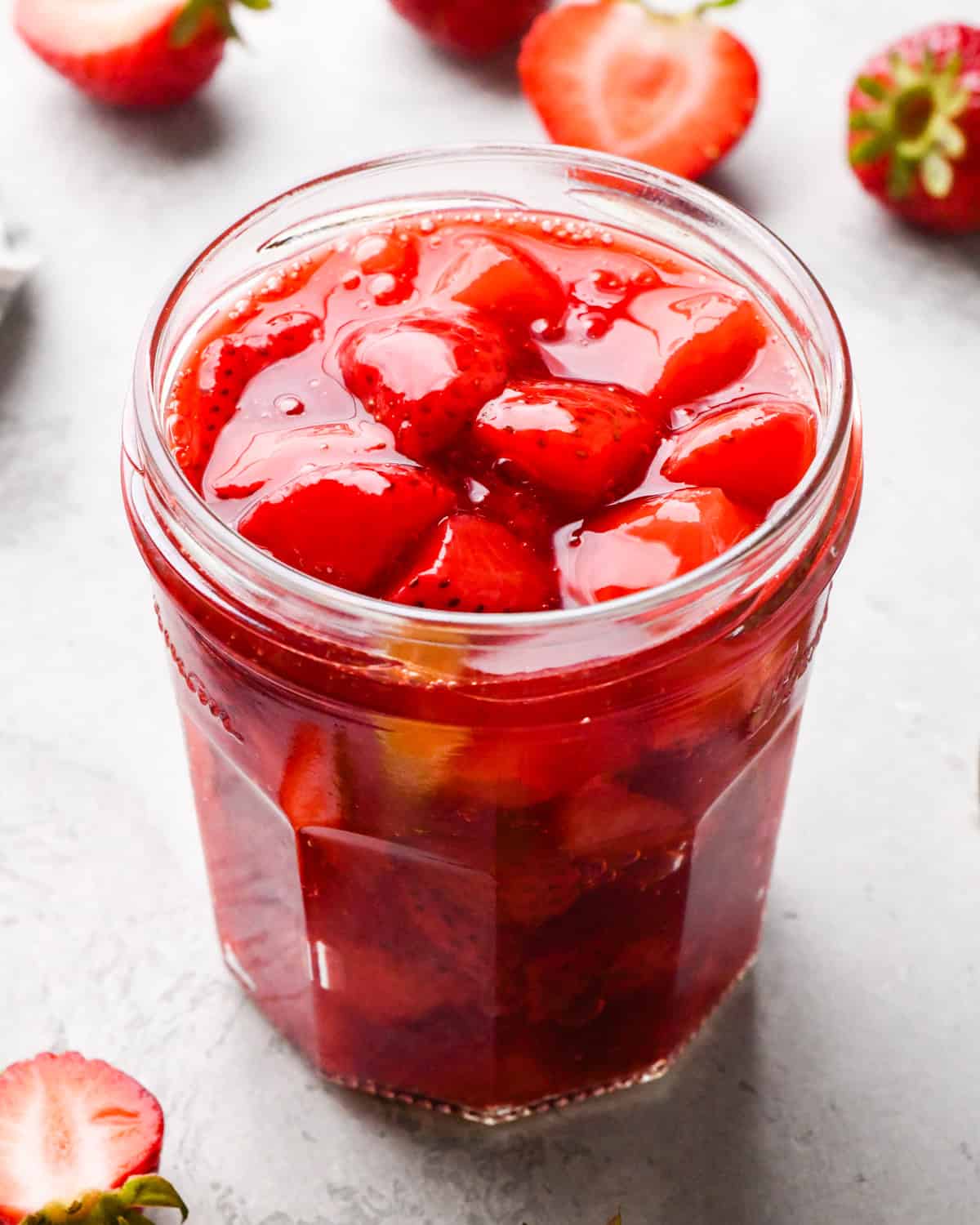 Overhead photo of a clear jar filled with bright red homemade strawberry sauce, with extra strawberries surrounding the jar on a light surface.