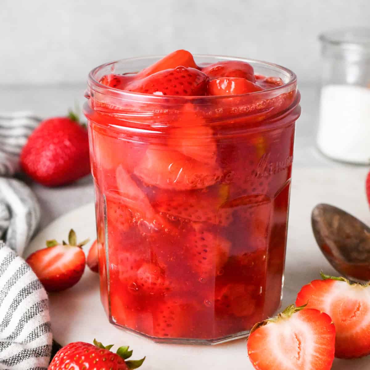 Overhead photo of a clear jar filled with bright red homemade strawberry sauce, with extra strawberries surrounding the jar on a light surface.