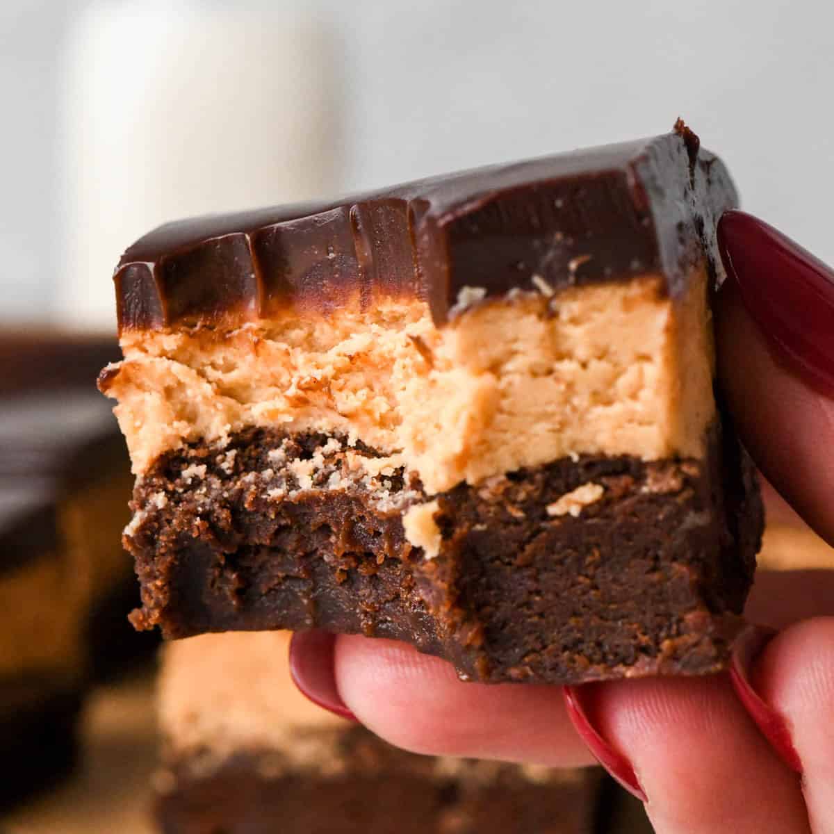 Up-close photo of a hand holding a buckeye brownie with a fudgy chocolate brownie base, creamy peanut butter layer, and smooth chocolate ganache. A bite has been taken out of the brownie.