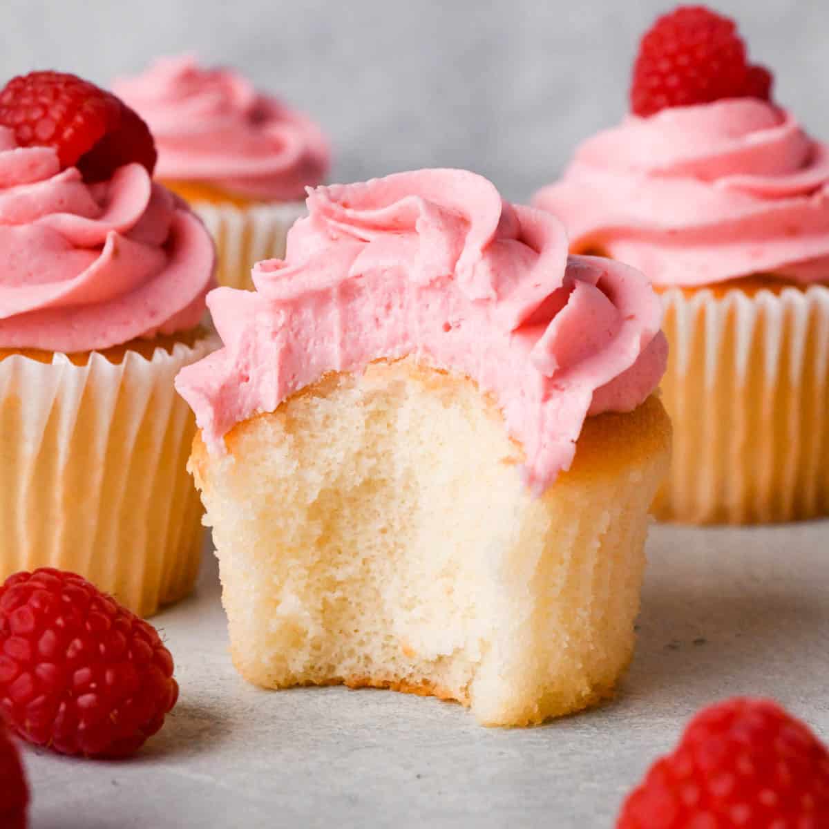 raspberry frosting on top of a vanilla cupcake with a bite taken out of it so the texture of the frosting is really visible, surrounded by more cupcakes topped with raspberry buttercream frosting
