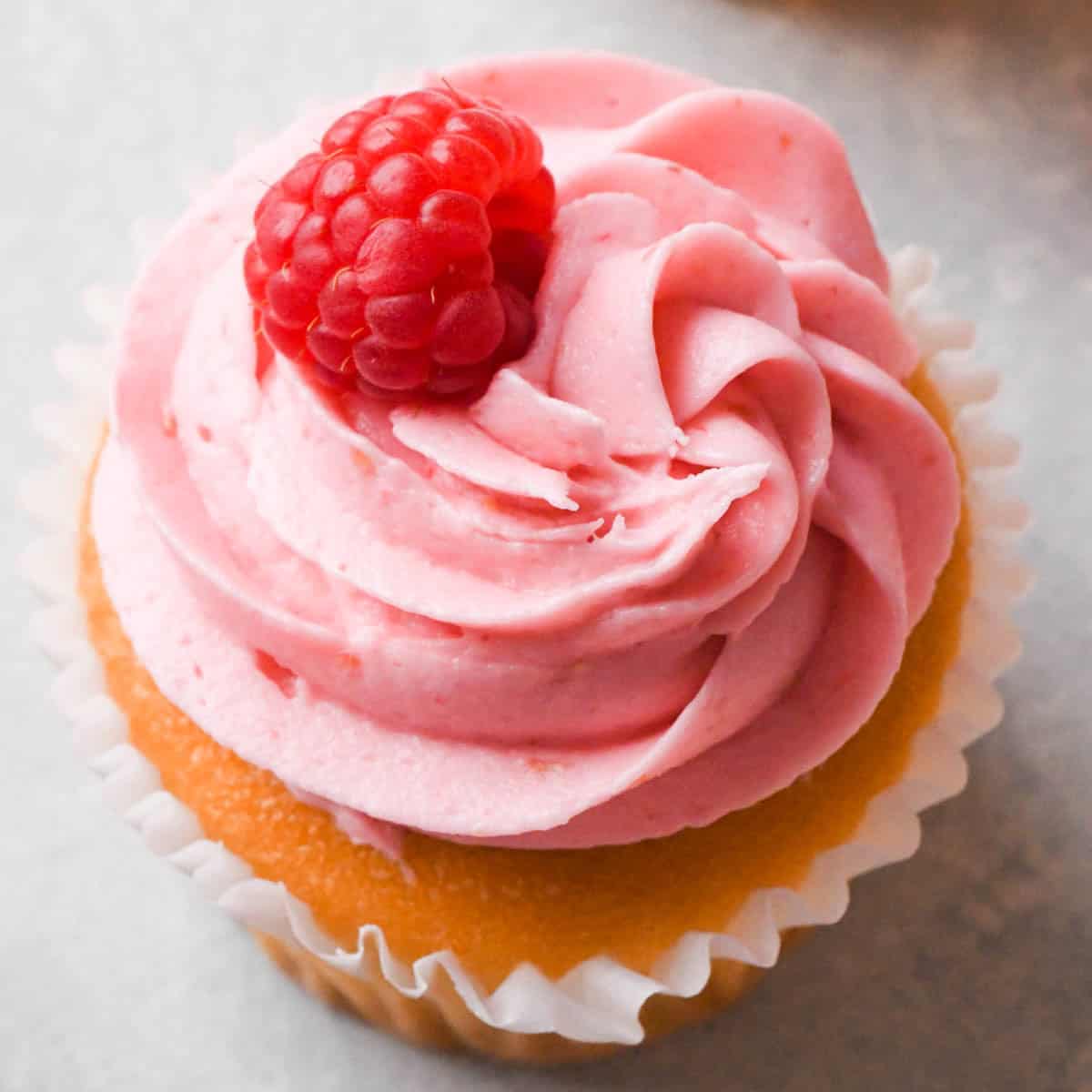 overhead view of raspberry frosting on top of a vanilla cupcake in a wrapper with a fresh raspberry on top