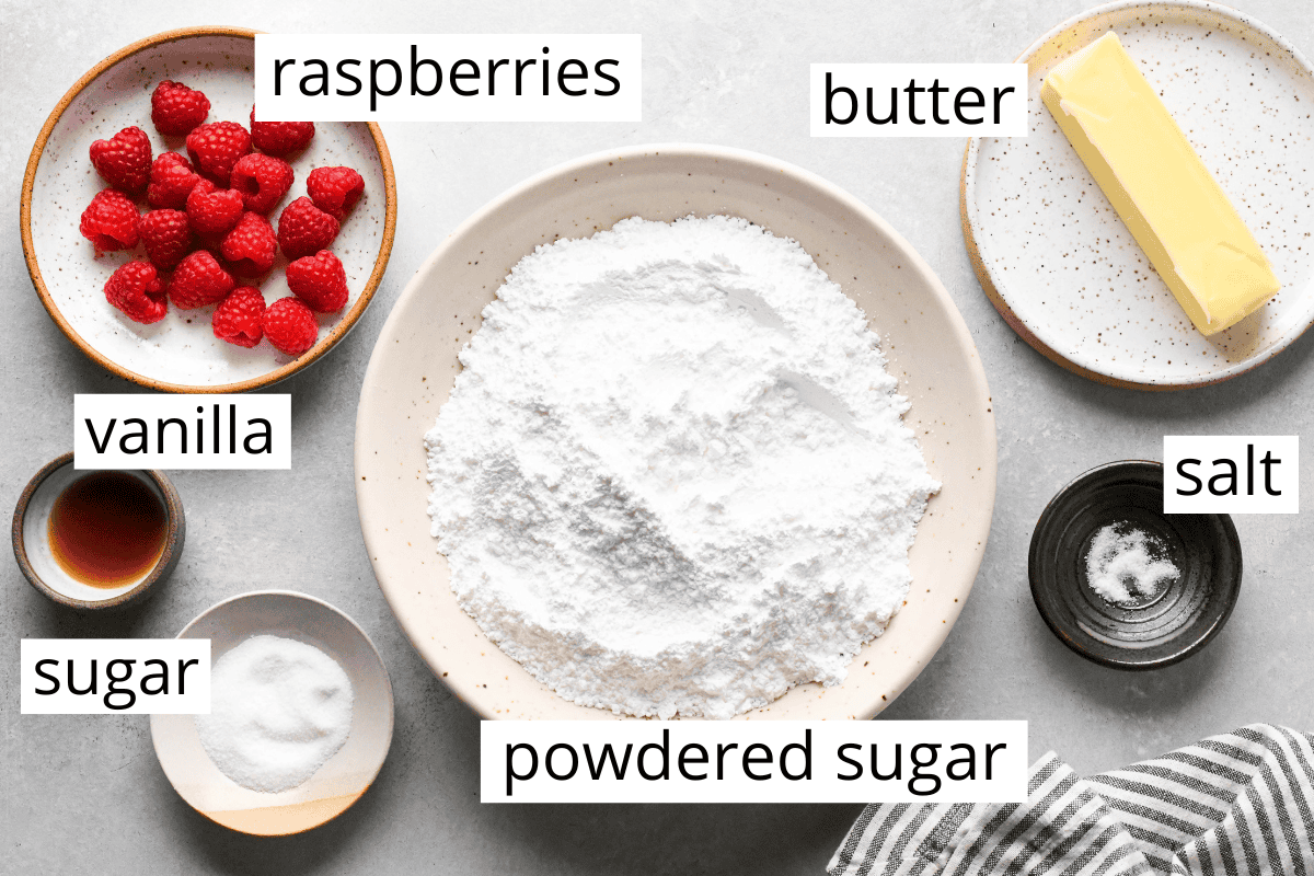 overhead photo of the ingredients in this raspberry frosting recipe all in bowls and labeled - butter, salt, powdered sugar, raspberries, vanilla and sugar