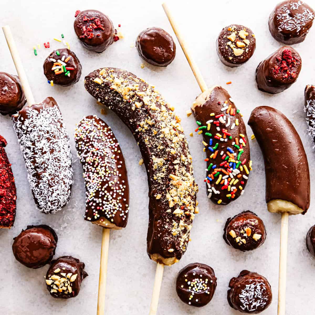 Overhead photo of chocolate dipped frozen bananas on sticks with assorted toppings including rainbow sprinkles, shredded coconut, crushed nuts, and freeze-dried berries, arranged on a light surface with small bowls of toppings nearby.