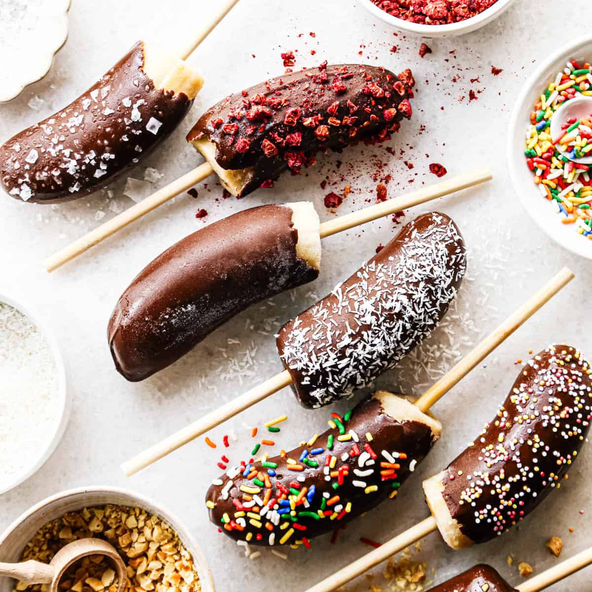 Overhead photo of chocolate dipped frozen bananas on sticks with assorted toppings including rainbow sprinkles, shredded coconut, crushed nuts, and freeze-dried berries, arranged on a light surface with small bowls of toppings nearby.