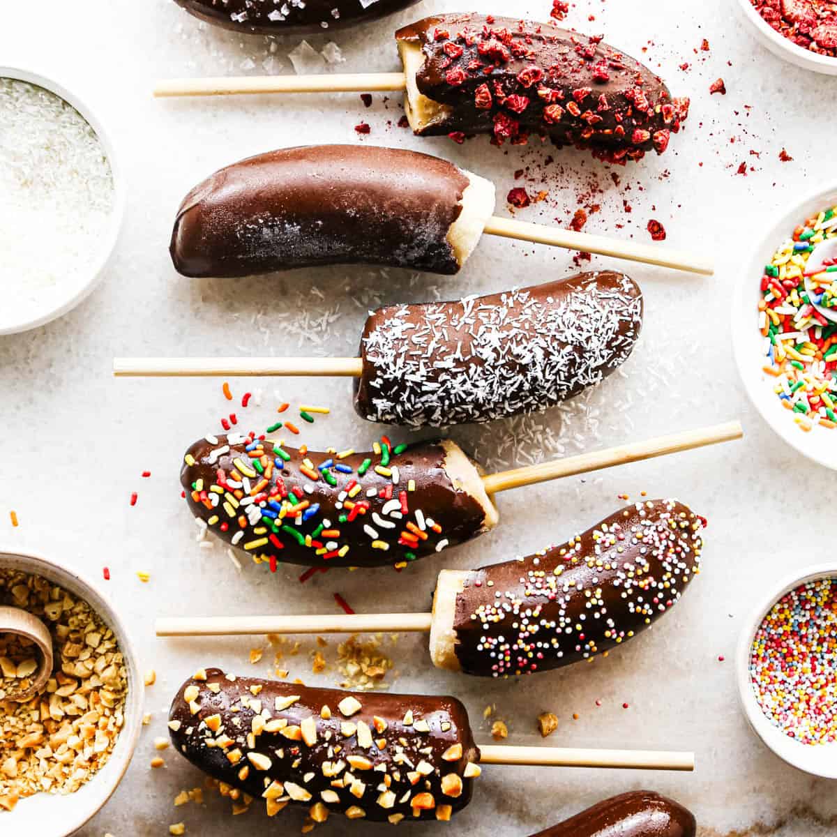 Making chocolate dipped frozen bananas: overhead photo of frozen banana halves arranged on a countertop after being dipped in melted chocolate and sprinkled with various toppings. Additional small bowls of toppings surround the baking sheet.