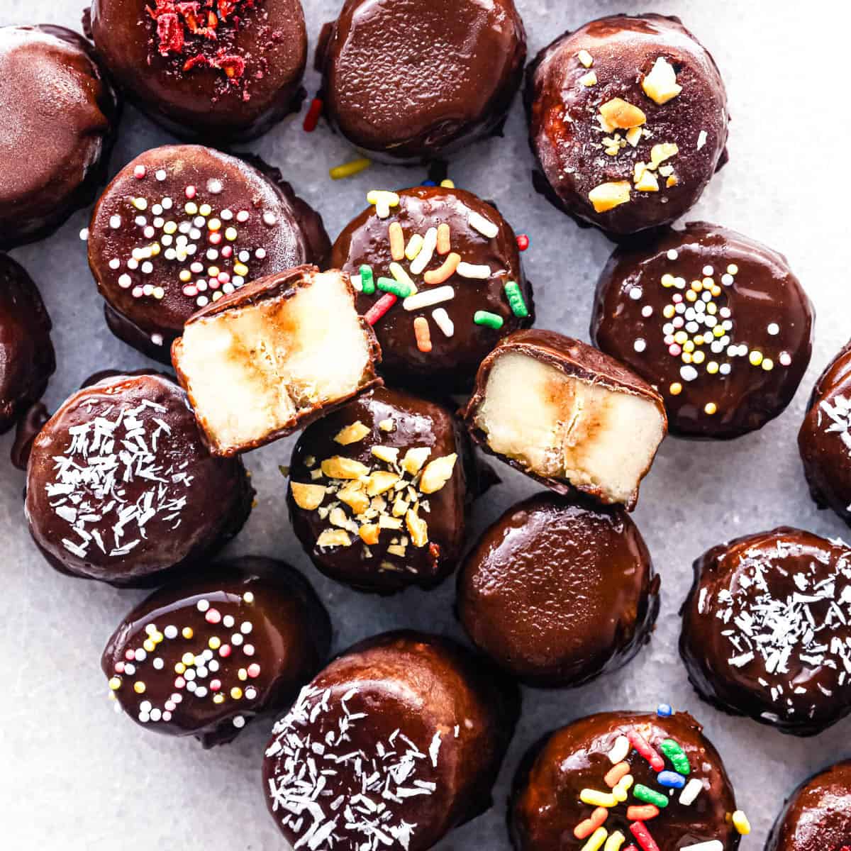 Up-close photo of a countertop full of chocolate dipped banana slices topped with various toppings. A bite is taken out of 2 of the banana slices to show the inside.