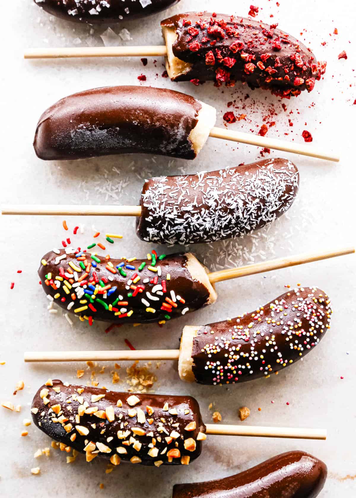 Making chocolate dipped frozen bananas: overhead photo of frozen banana halves arranged on a countertop after being dipped in melted chocolate and sprinkled with various toppings.