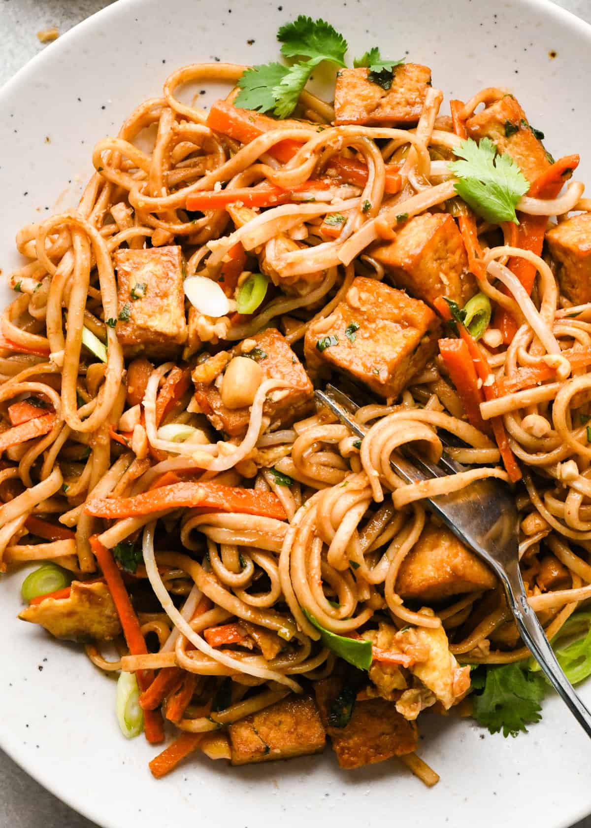 Close-up photo of a plate of homemade pad Thai with rice noodles, crispy tofu, carrots, and scallions, garnished with cilantro, crushed peanuts, and lime wedges. The meal is being eaten with a fork.