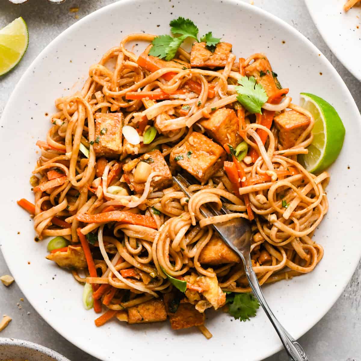 Overhead photo of a plate of homemade pad Thai with rice noodles, crispy tofu, carrots, and scallions, garnished with cilantro, crushed peanuts, and lime wedges. The meal is being eaten with a fork.