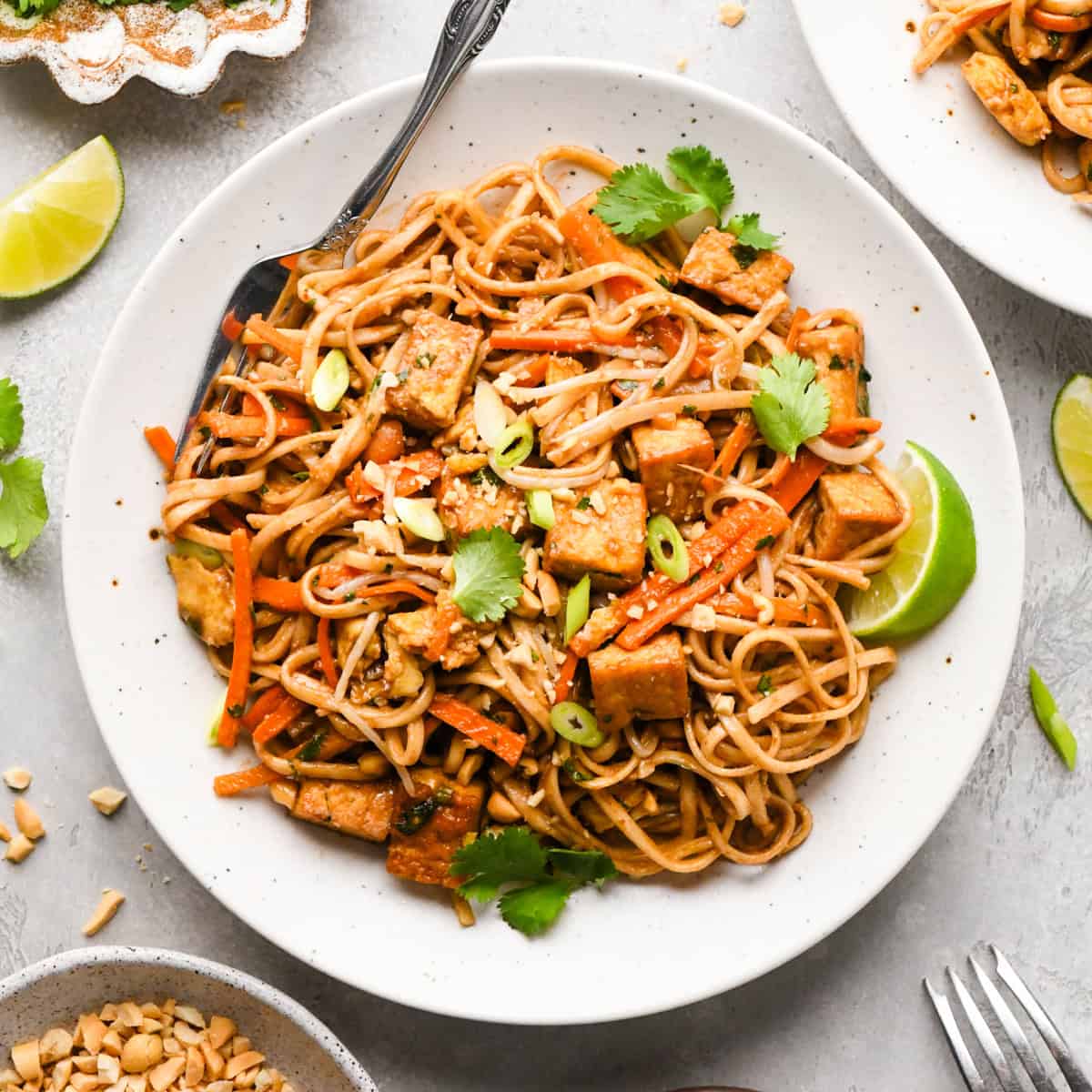 Overhead photo of a plate of homemade pad Thai with rice noodles, crispy tofu, carrots, and scallions, garnished with cilantro, crushed peanuts, and lime wedges. A fork is resting on the white plate.