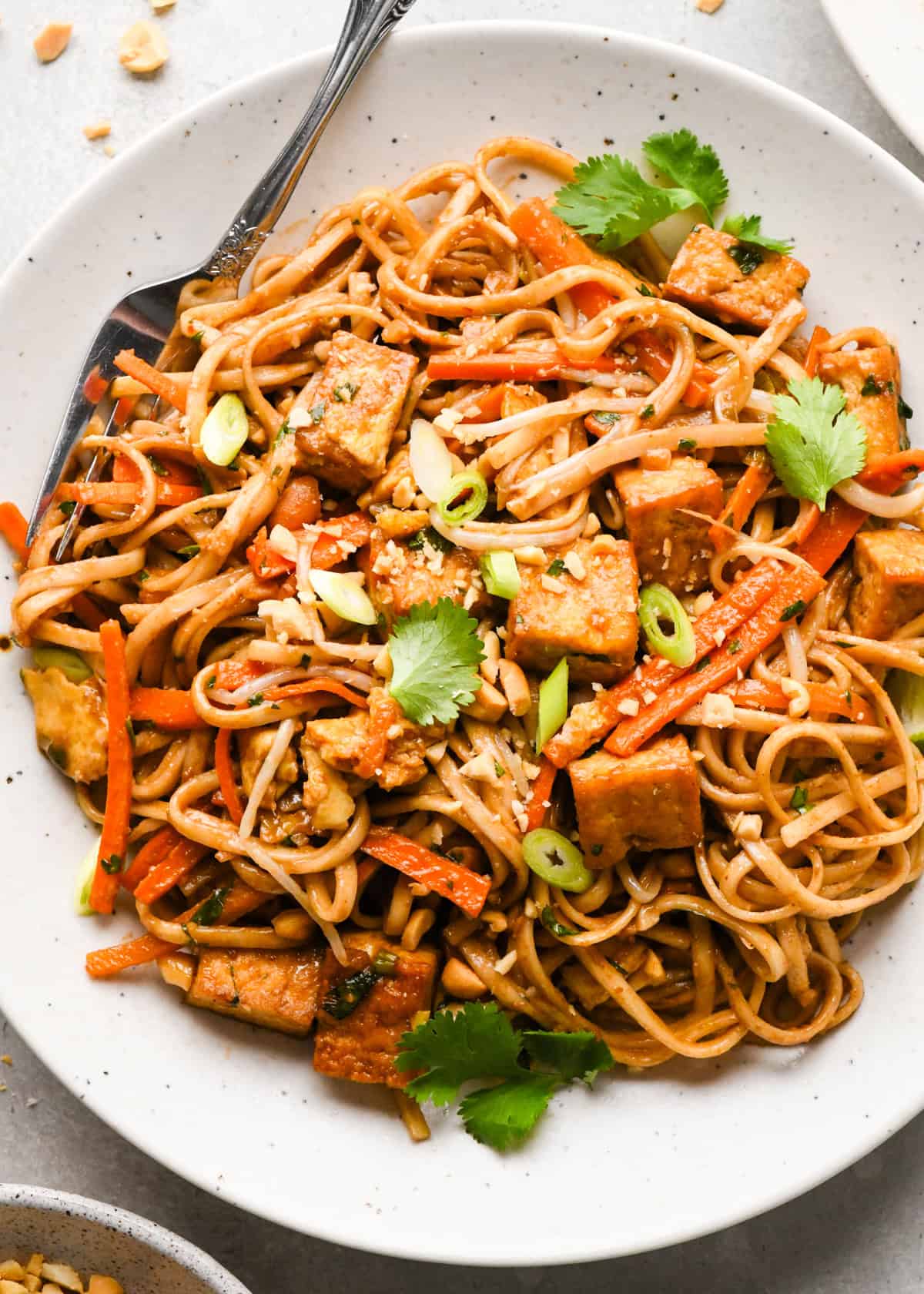 Overhead photo of a plate of homemade pad Thai with rice noodles, crispy tofu, carrots, and scallions, garnished with cilantro, crushed peanuts, and lime wedges. A fork is resting on the white plate.