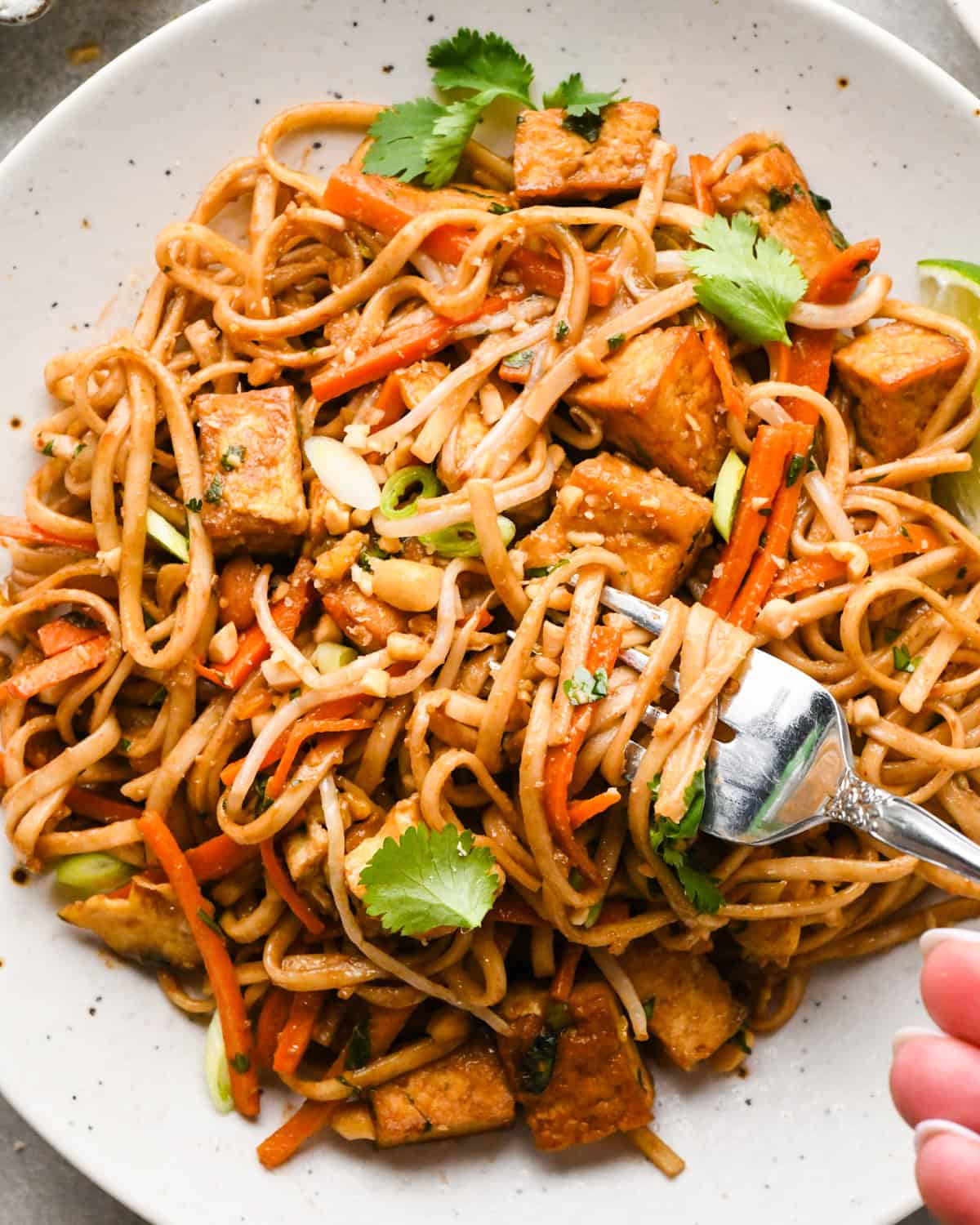 Close-up photo of a plate of homemade pad Thai with rice noodles, crispy tofu, carrots, and scallions, garnished with cilantro, crushed peanuts, and lime wedges. The meal is being eaten with a fork.
