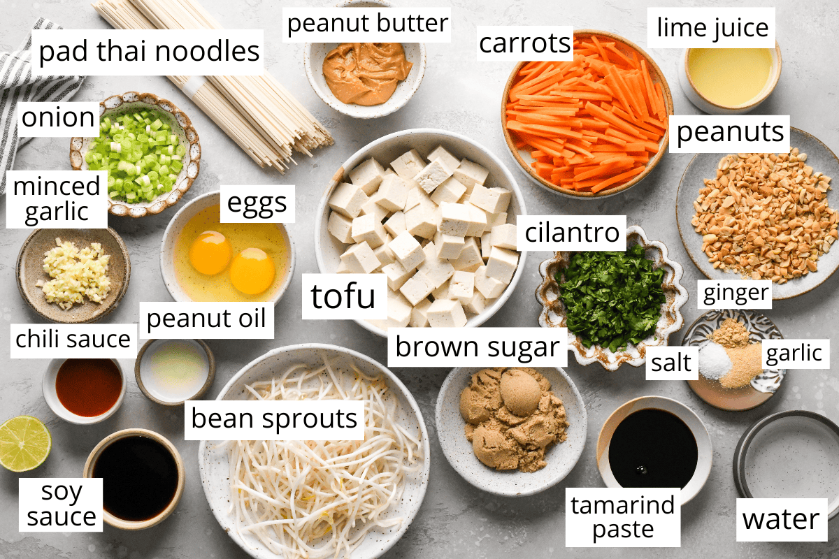 Overhead view of labeled pad thai ingredients arranged in bowls, including rice noodles, tofu, eggs, carrots, bean sprouts, peanuts, cilantro, onion, garlic, ginger, brown sugar, tamarind paste, soy sauce, chili sauce, peanut butter, peanut oil, lime juice, salt, and water.