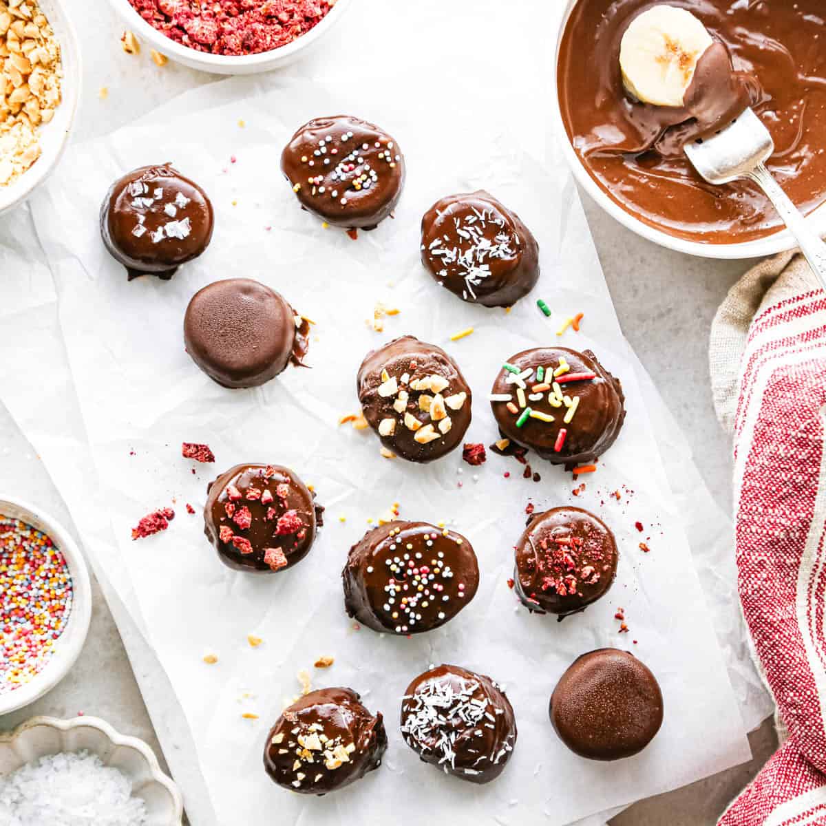Making chocolate dipped frozen bananas: overhead photo of 12 single banana slices arranged on a sheet of parchment paper after being dipped in chocolate and sprinkled with various toppings. Additional small bowls of toppings surround the bananas on a countertop.