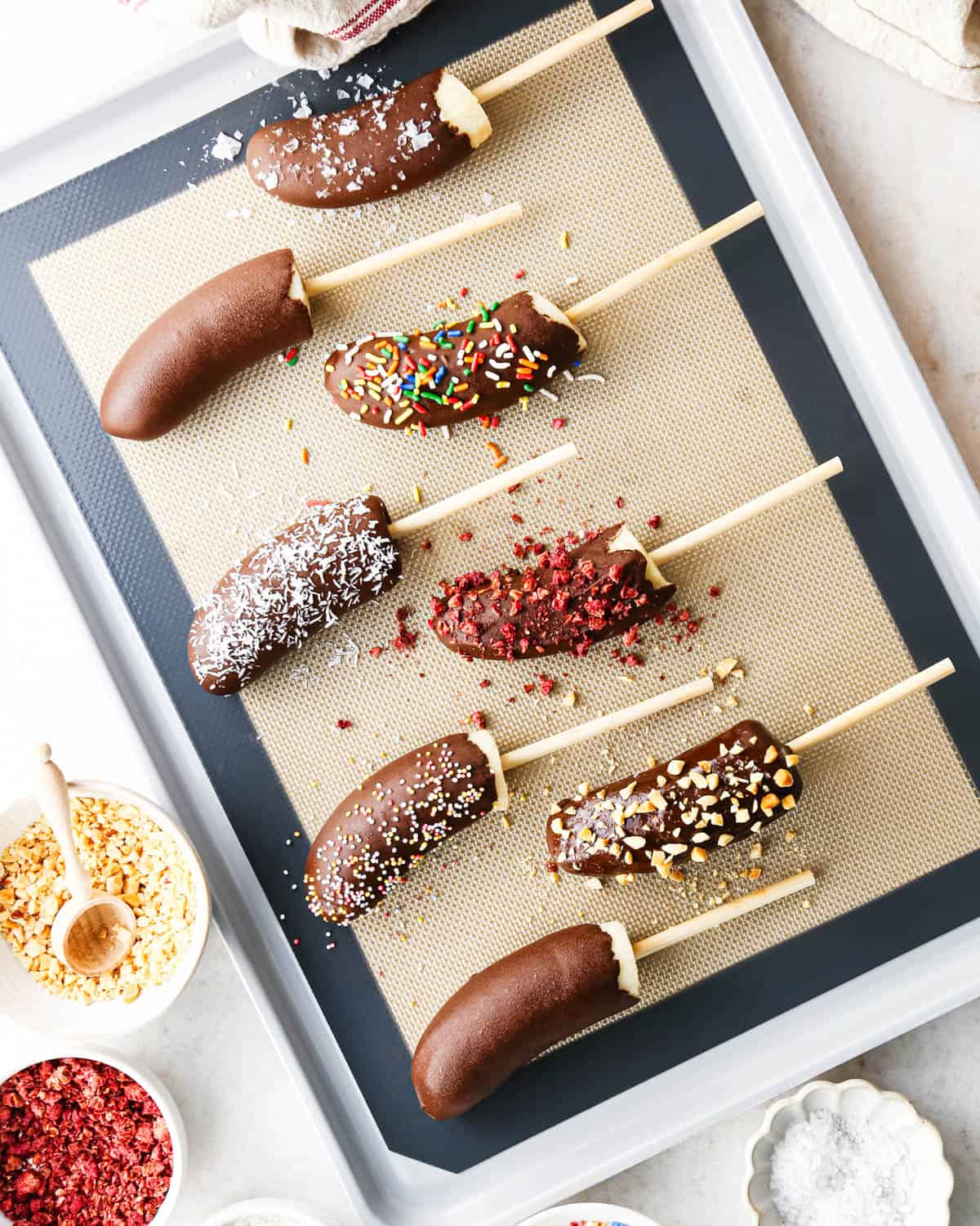 Making chocolate dipped frozen bananas: overhead photo of frozen banana halves arranged on a baking sheet after being dipped in melted chocolate and sprinkled with various toppings. Additional small bowls of toppings surround the baking sheet.