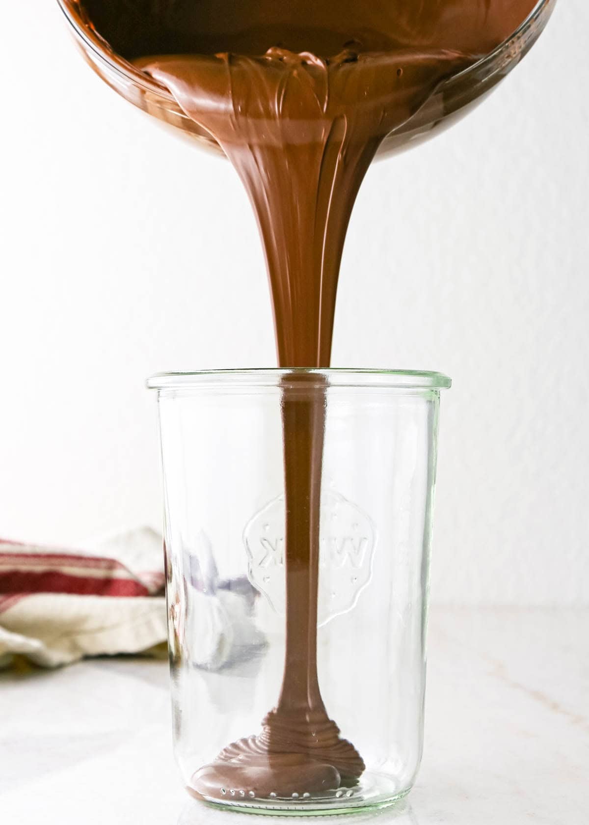 Making chocolate dipped frozen bananas: close-up photo of melted chocolate being poured into a tall clear glass on a countertop.