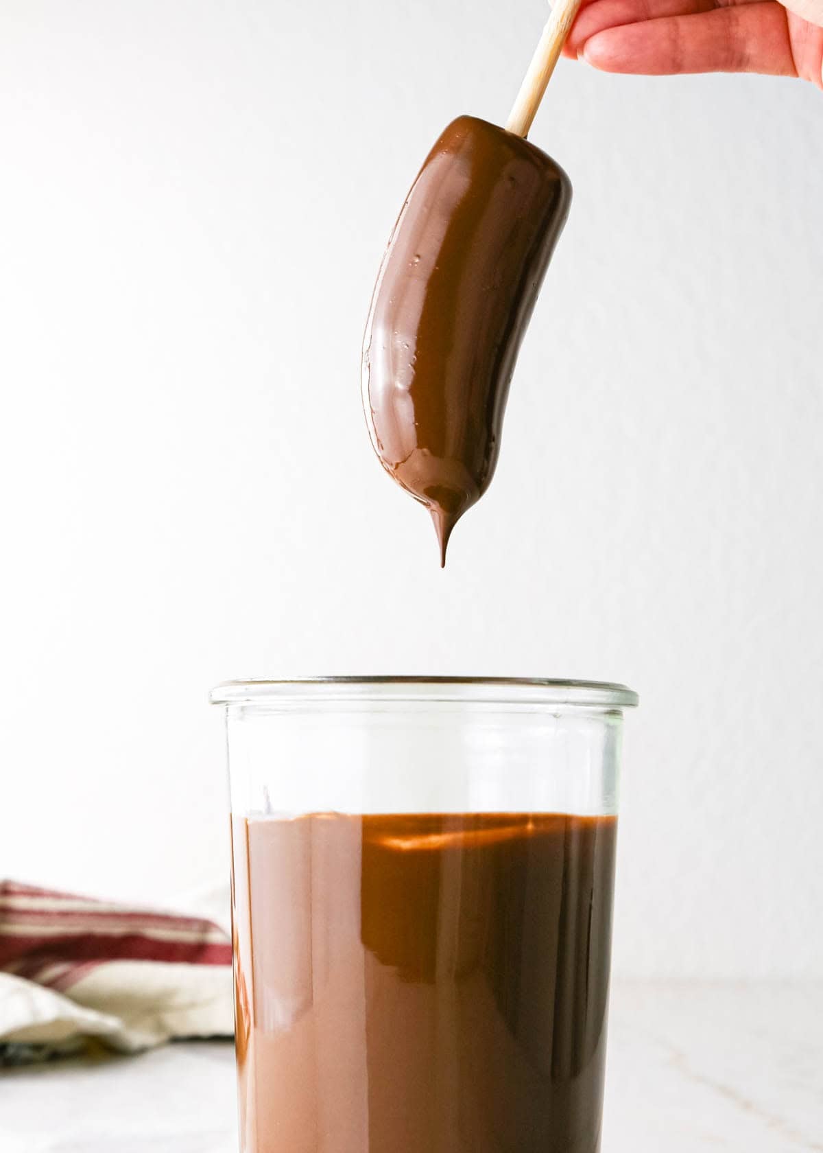 Making chocolate dipped frozen bananas: close-up photo of a frozen half banana on a candy stick after being dipped in melted chocolate in a tall clear glass.