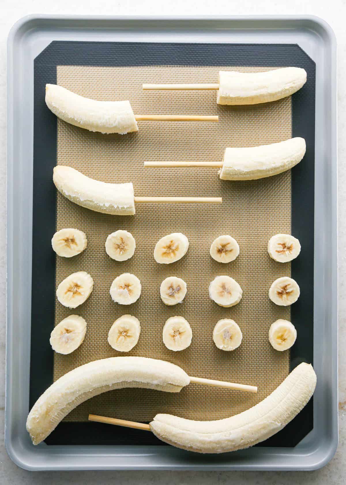 Making chocolate dipped frozen bananas: overhead photo of a metal baking sheet lined with a silicone baking mat and filled with frozen bananas. Some bananas are whole with candy sticks in them, some are cut in half with candy sticks in them, and in the center there are 3 rows of single banana slices.