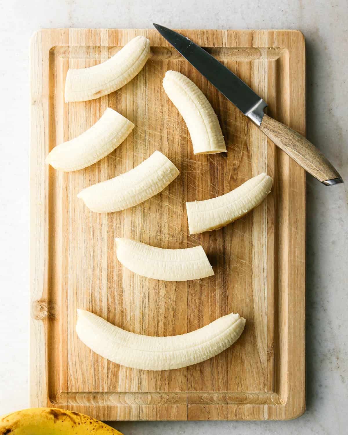 Overhead photo of a wooden cutting board full of halved bananas ready for dipping in chocolate. A knife is sitting on the cutting board.