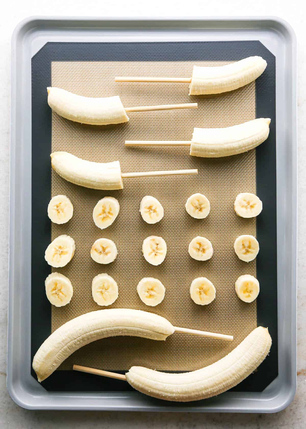 Making chocolate dipped frozen bananas: overhead photo of a metal baking sheet lined with a silicone baking mat and filled with prepared bananas. Some bananas are whole with candy sticks in them, some are cut in half with candy sticks in them, and in the center there are 3 rows of single banana slices.