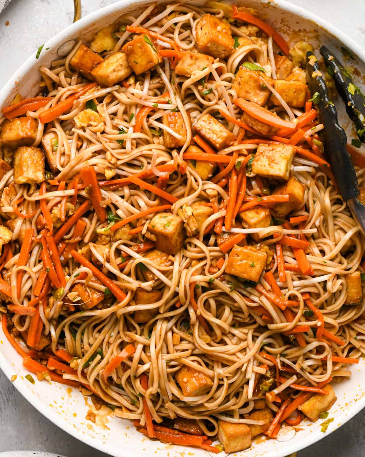 Making Pad Thai: overhead photo of cooked noodles, bean sprouts, cilantro and sauce added in with the tofu and vegetable mixture in a skillet and tossed together with tongs.