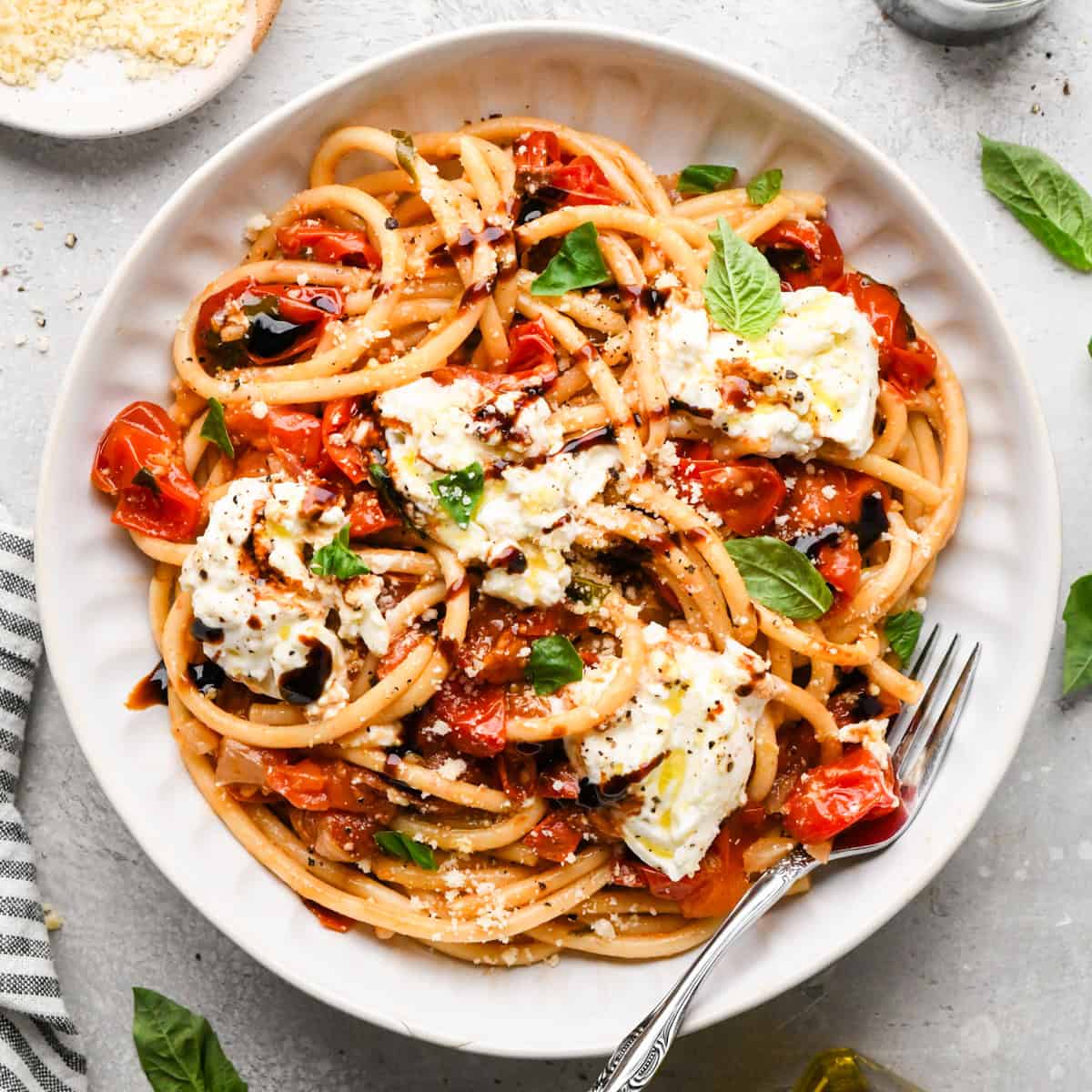 Overhead photo of a white bowl of pasta tossed with roasted tomatoes and fresh basil, topped with creamy burrata cheese and a drizzle of balsamic glaze. Served with a fork.