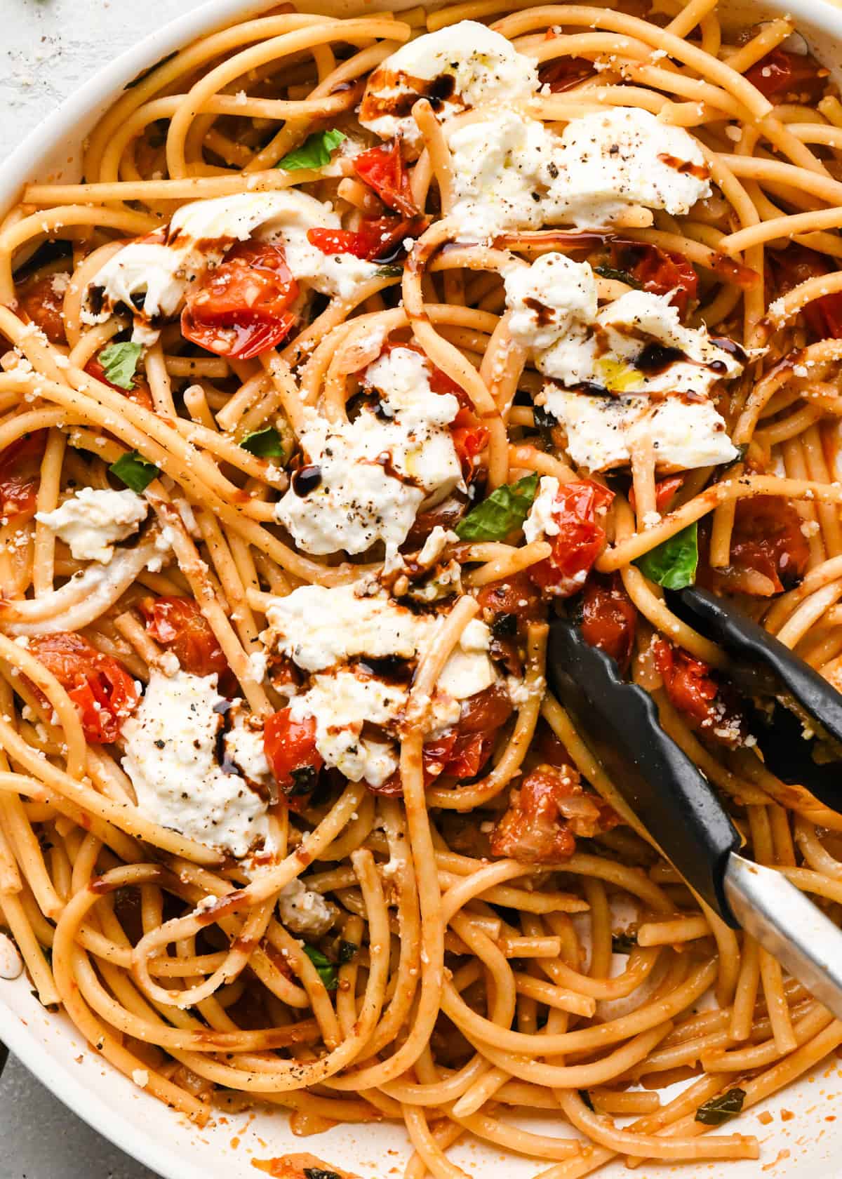Making pasta with burrata cheese: close-up photo of completed pasta meal with burrata cheese, roasted tomatoes and basil, and balsamic vinegar being served with tongs.
